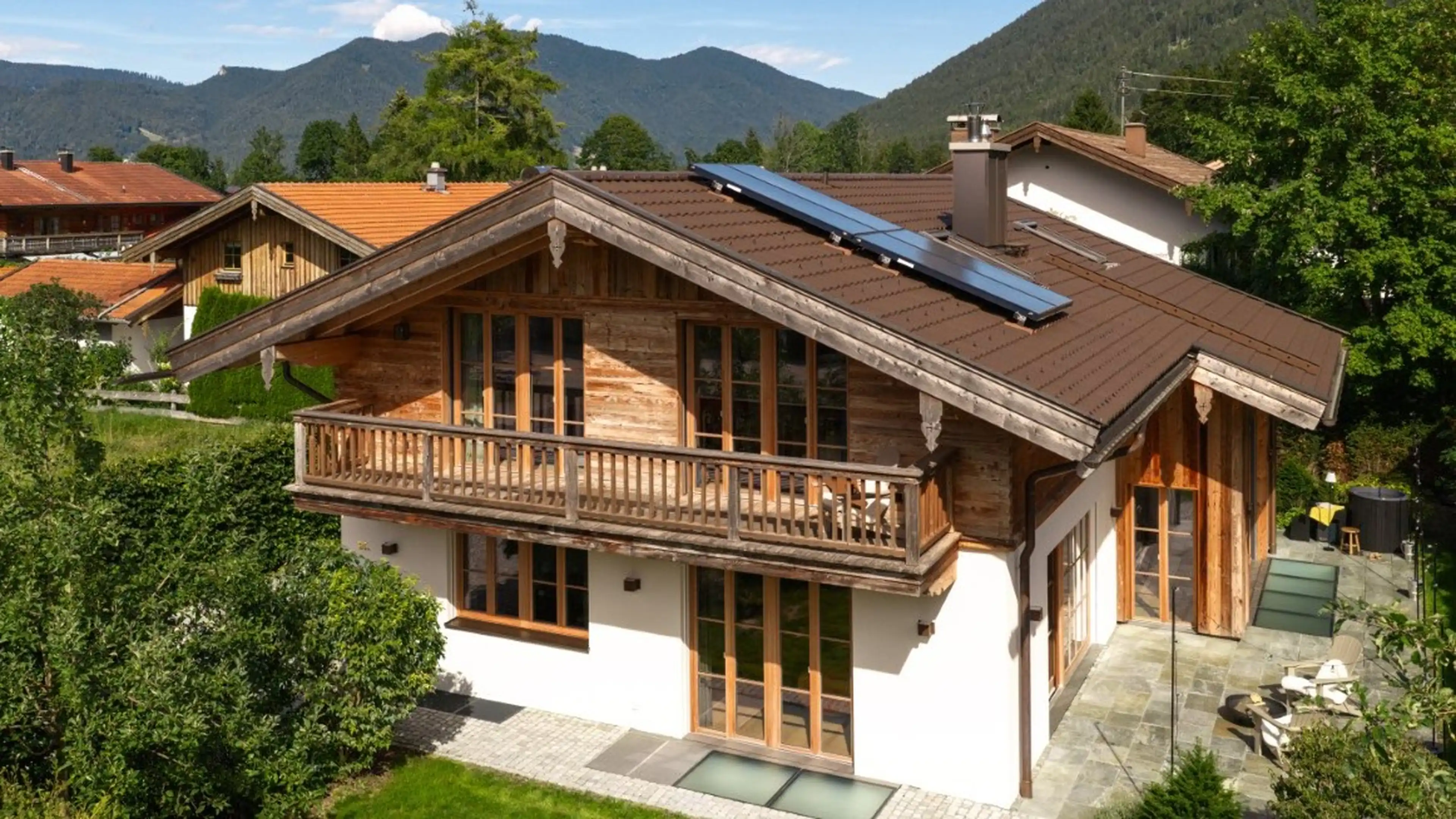 Luftaufnahme eines zweistöckigen Landhauses mit weiß verputztem Erdgeschoss, dunkler Holzfassade im Obergeschoss, braunem Ziegeldach mit Solarpaneelen und umlaufendem Holzbalkon, umgeben von Bäumen, Terrasse und Bergpanorama im Hintergrund.