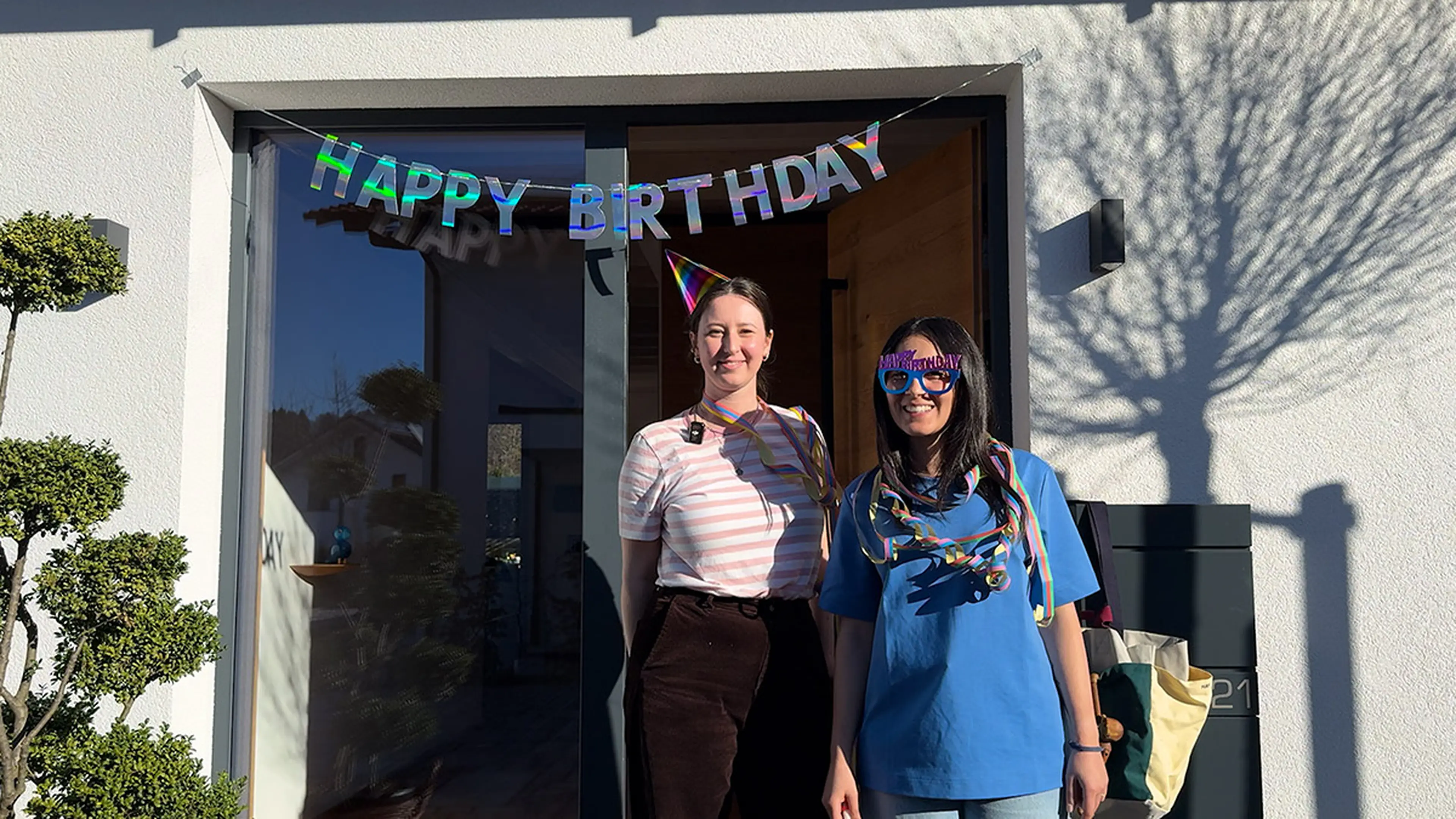 Steffi und Tugce von der Traumhausverlosung stehen vor der Eingangstür des Traumhauses am Chiemsee über der eine bunte Happy Birthday Girlande hängt. Geschmückt sind die beiden Frauen mit einem Partyhut, einer Partybrille und Luftschlangen um den Hals