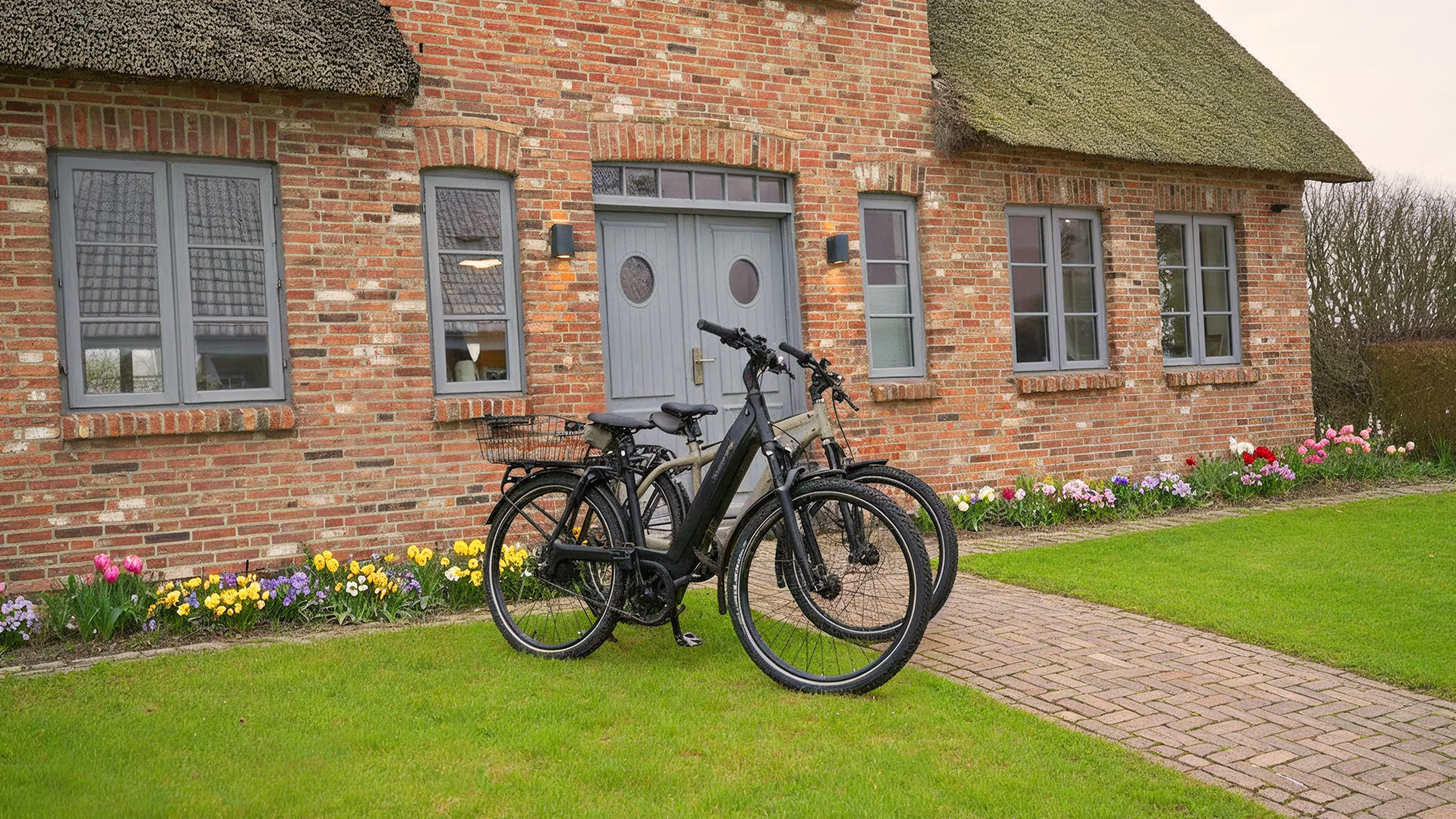 2 E-Bikes in grau, seitlich fotografiert vor dem Traumhaus auf Föhr umgeben von grüner Rasenfläche und einem frühlingshaften Vorgarten