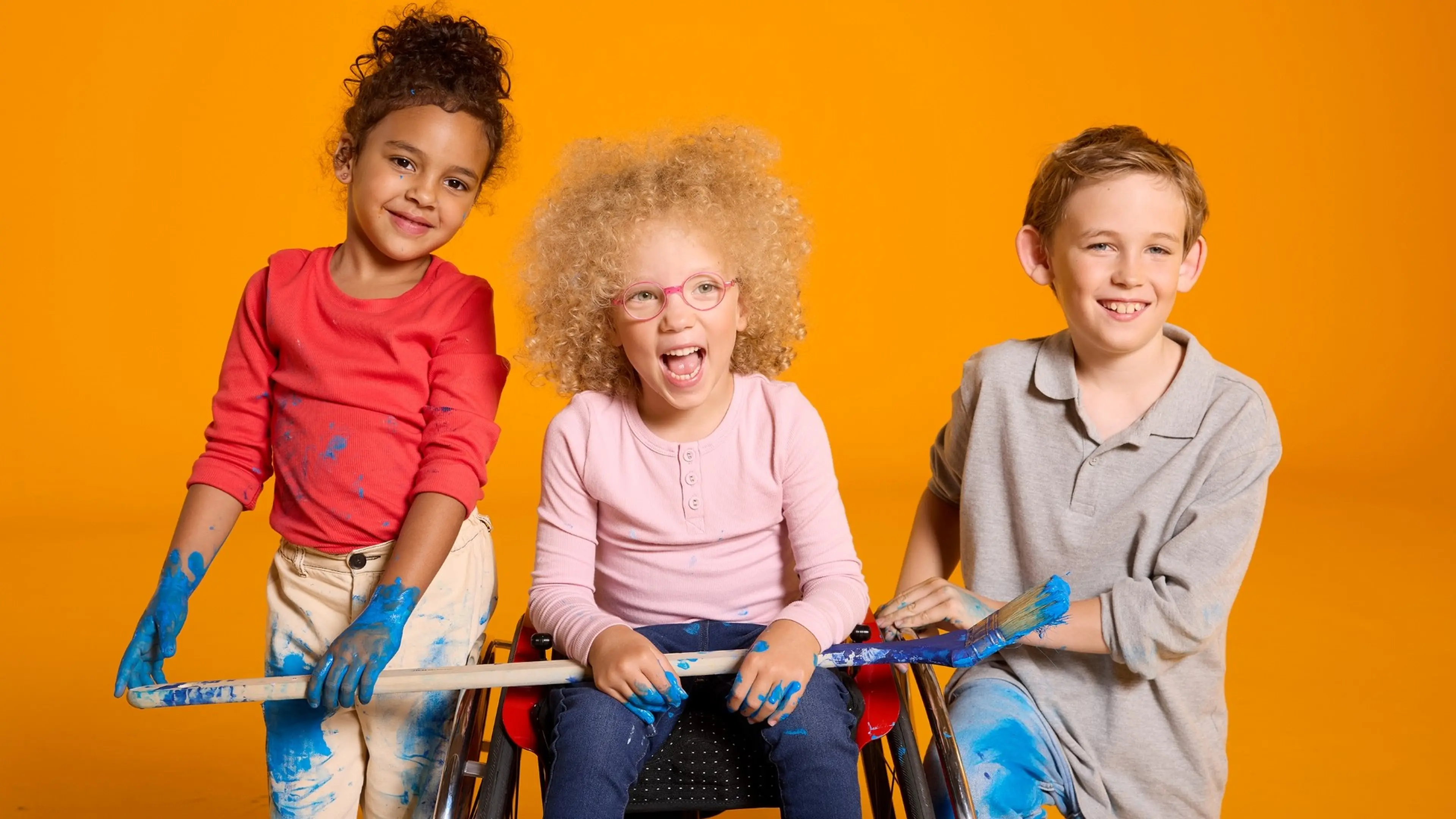 3 lachende Kindern, die mit blauer Farbe beschmiert sind und einen großen Pinsel halten