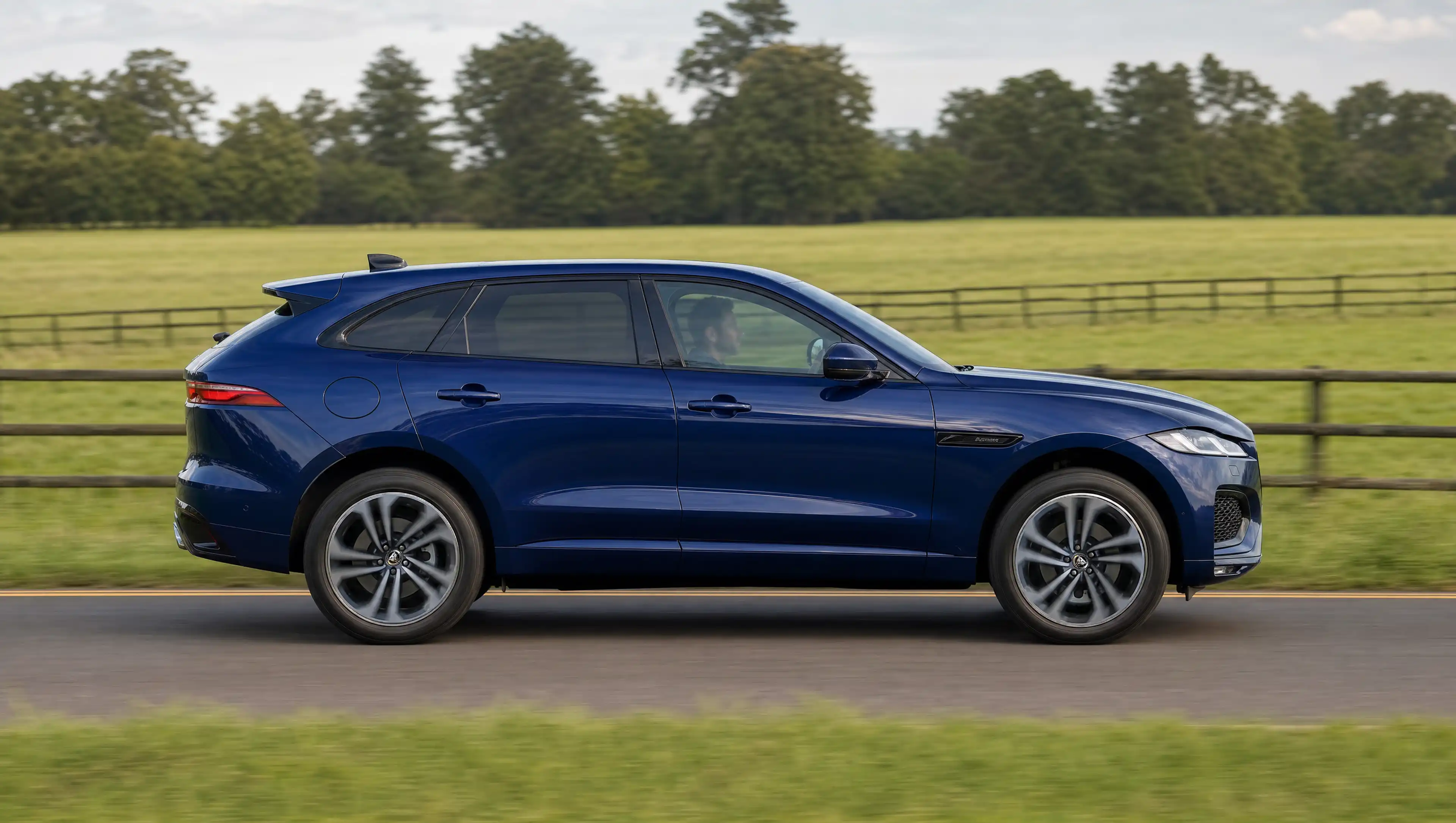Ein blauer Jaguar F-Pace seitlich fotografiert auf einer Landstraße umgeben von Pferdekoppeln