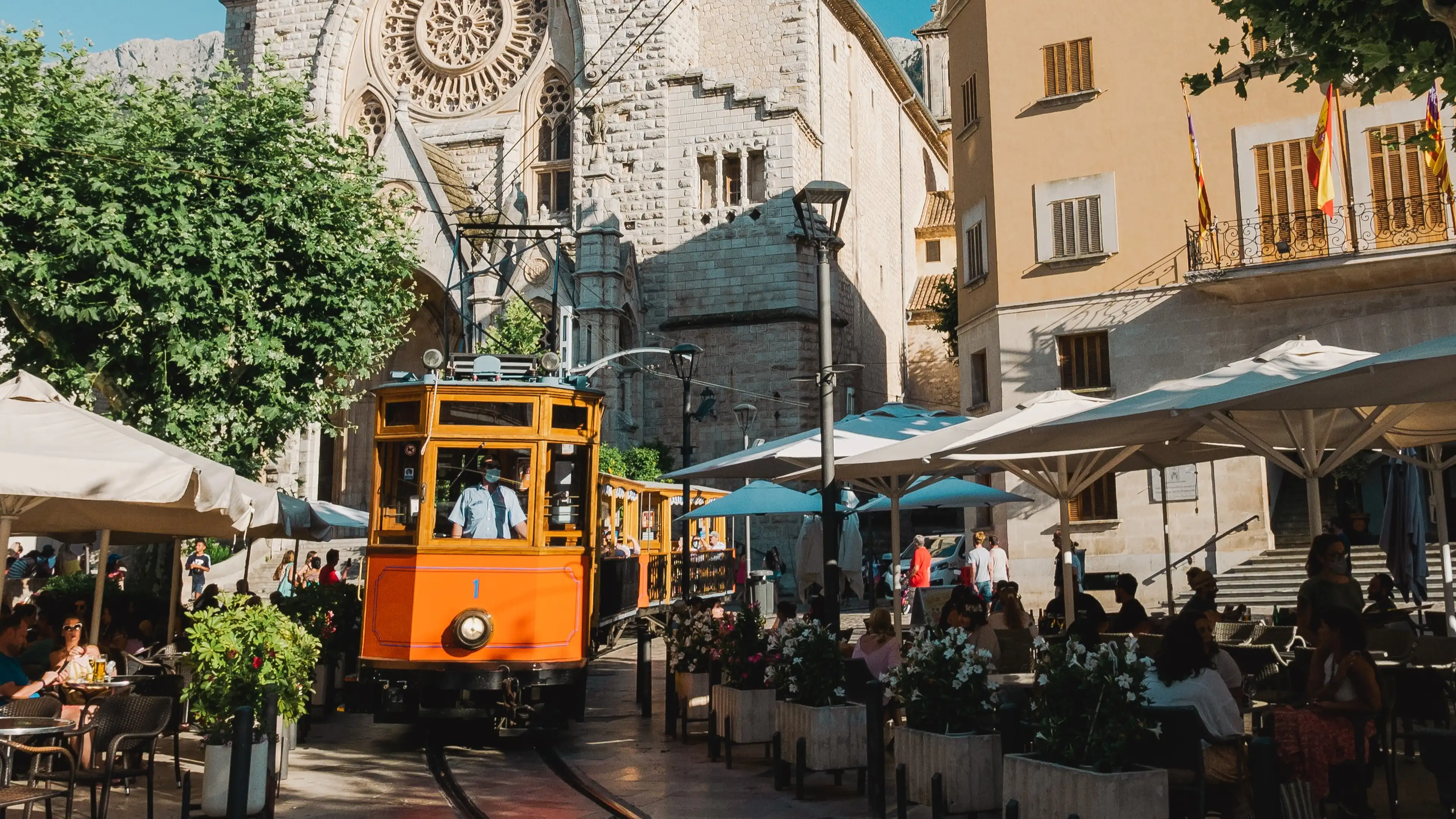 Historische Straßenbahn von Sóller fährt durch die belebte Altstadt vor der beeindruckenden Kirche Sant Bartomeu auf Mallorca, umgeben von Cafés und mediterranem Flair.