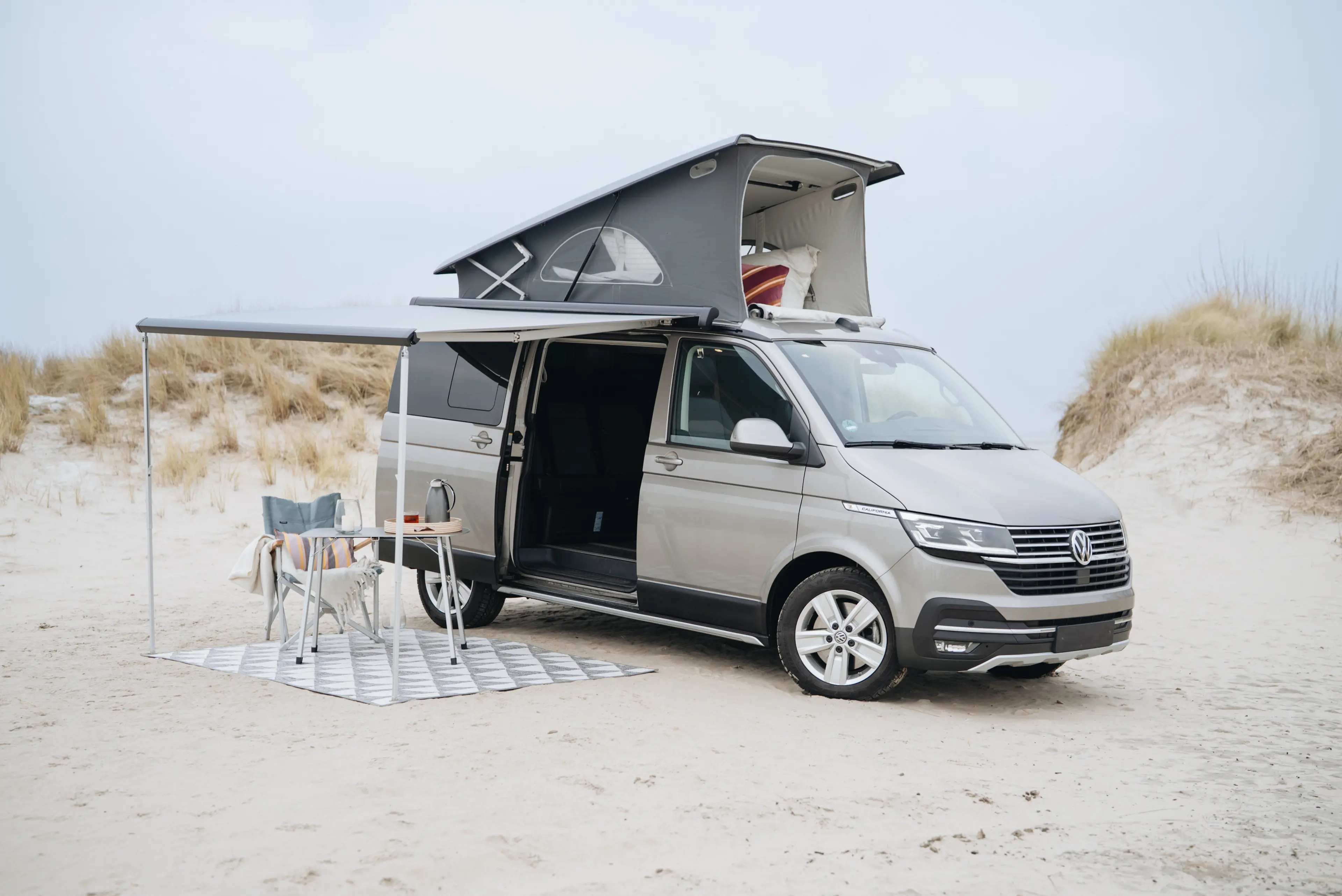 Der VW California mit ausgeklappter Schlafkabine und ausgefahrenem Dach unter welchem Campingstuhl und Tisch stehen, direkt am Strand auf Föhr