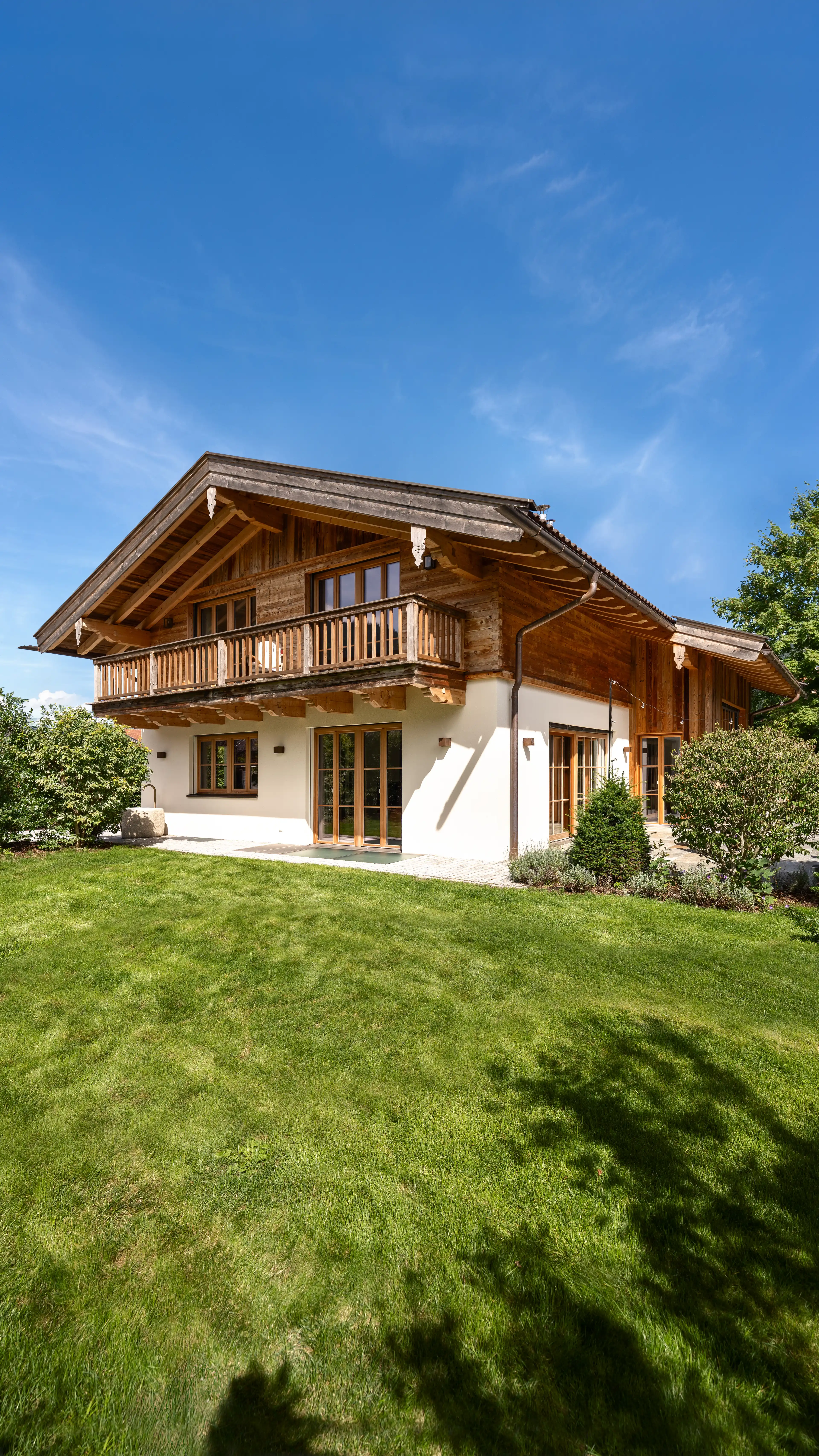 Frontansicht eines zweistöckigen Landhauses mit dunkler Holzfassade und auskragendem Giebeldach im Obergeschoss, weiß verputztem Erdgeschoss mit Holzfenstern und Terrassentüren, umgeben von grünem Rasen, Hecken und Bäumen unter blauem Himmel.