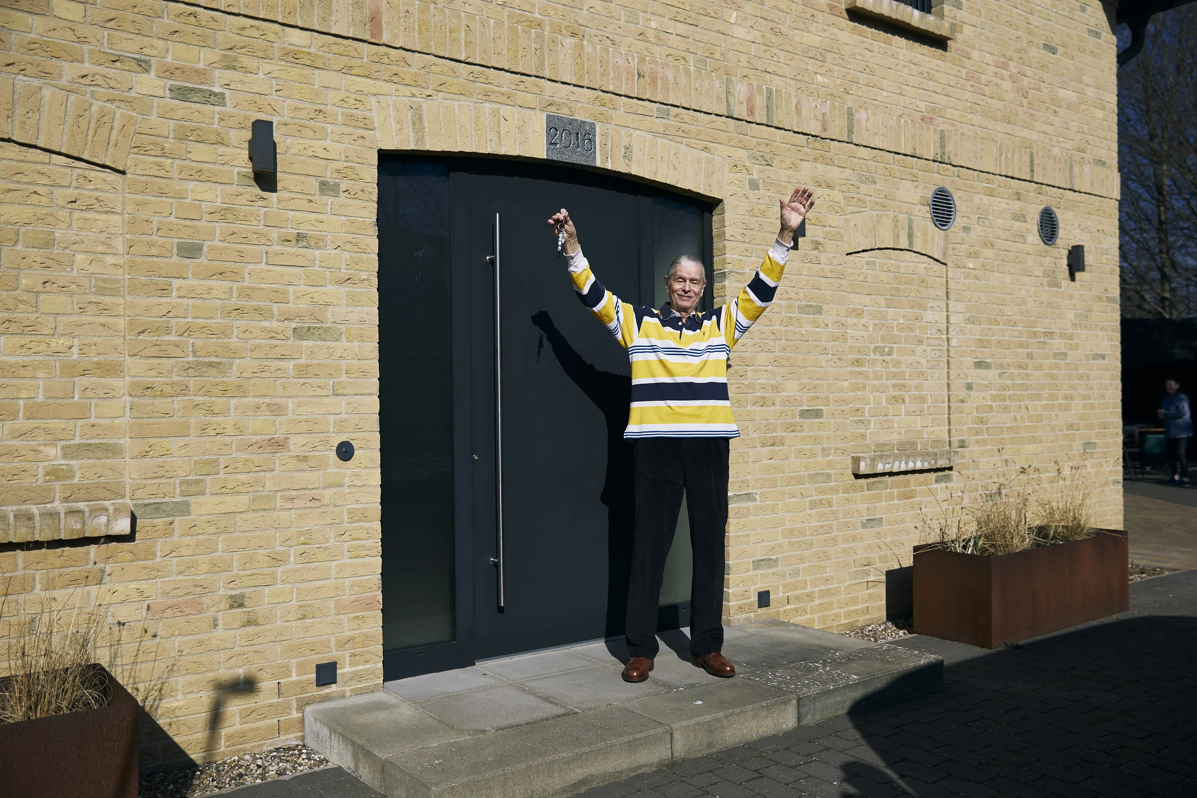 Mit ausgestreckten Armen und voller Freude steht Jürgen der Gewinner des Traumhauses mit dem Schlüssel in der Hand vor der Eingangstür seines neuen Eigenheims