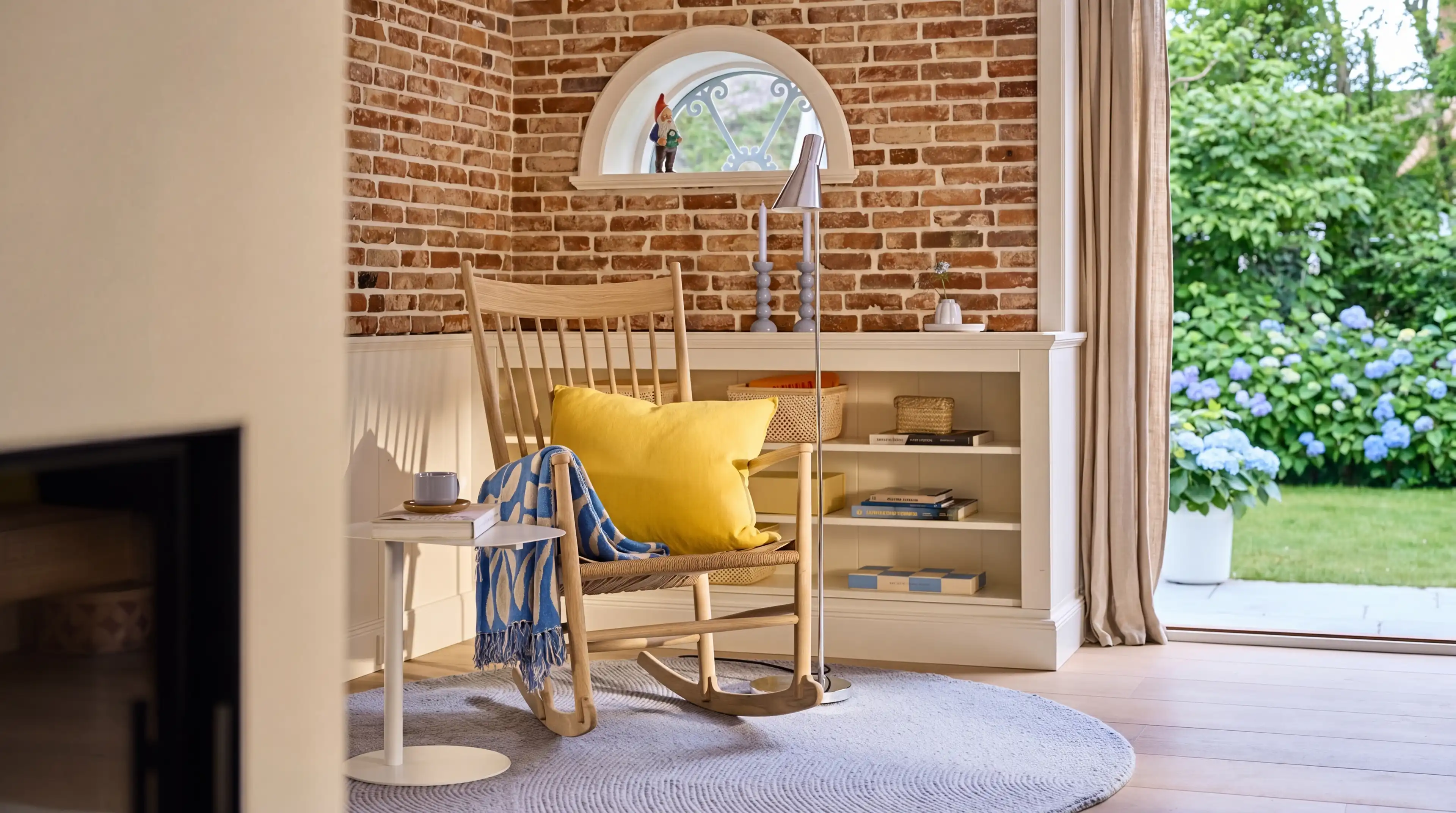 Eine Leseecke links neben der Terrassentür mit halbhohen weißen Bücherregalen, einem Schaukelstuhl aus Holz mit gelbem Kissen und einer backsteinfarbenden Wand mit kleinem weißem Fenster im Hintergrund.