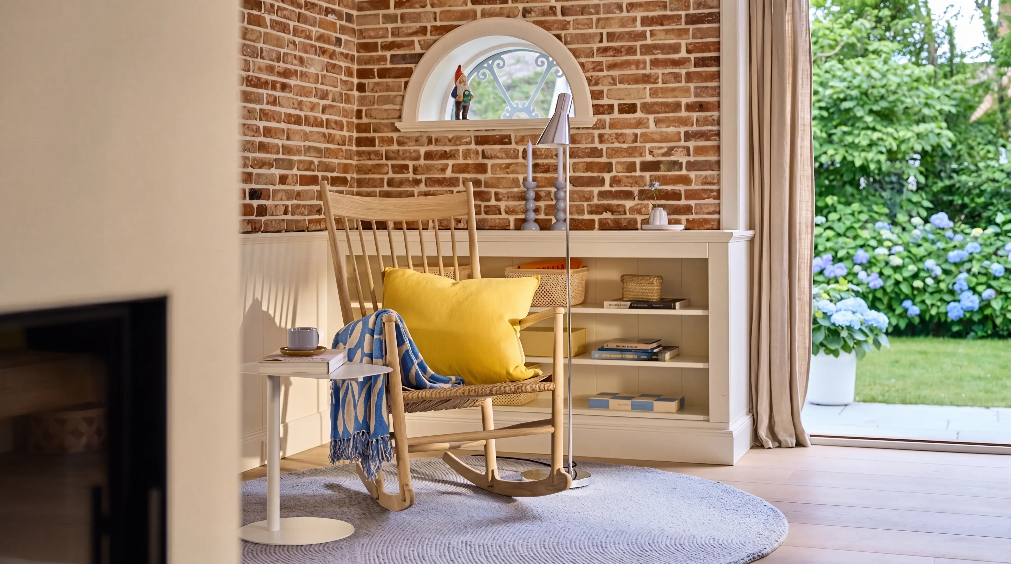 Eine Leseecke links neben der Terrassentür mit halbhohen weißen Bücherregalen, einem Schaukelstuhl aus Holz mit gelbem Kissen und einer backsteinfarbenden Wand mit kleinem weißem Fenster im Hintergrund.