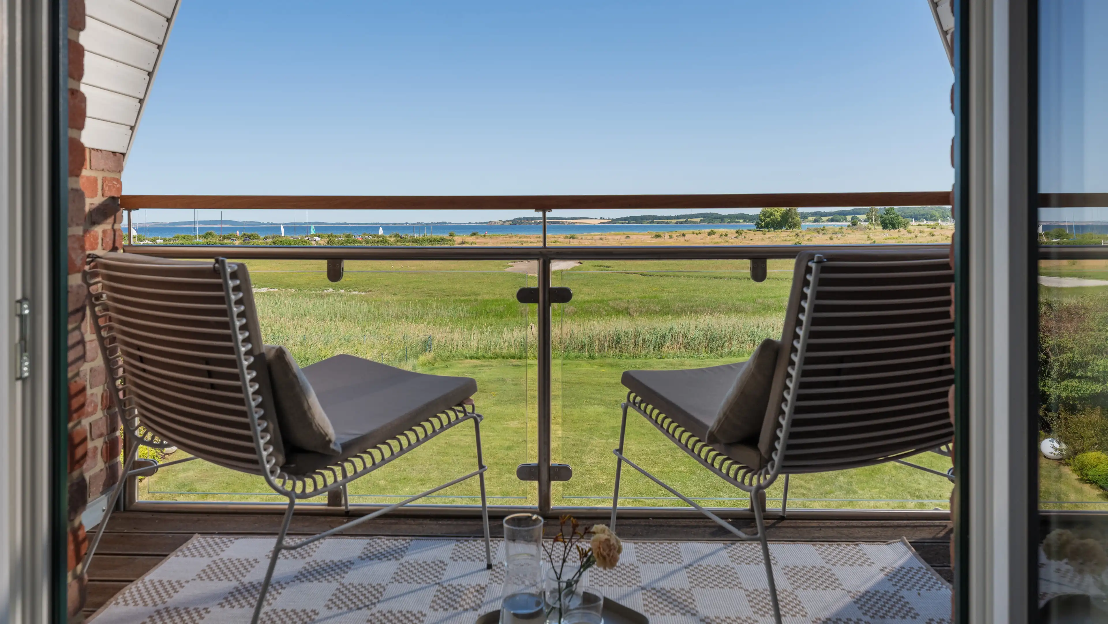 Blick vom Balkon über zwei Sitzstühle hinweg auf Wiese, Dünen und die Ostsee in der Ferne.