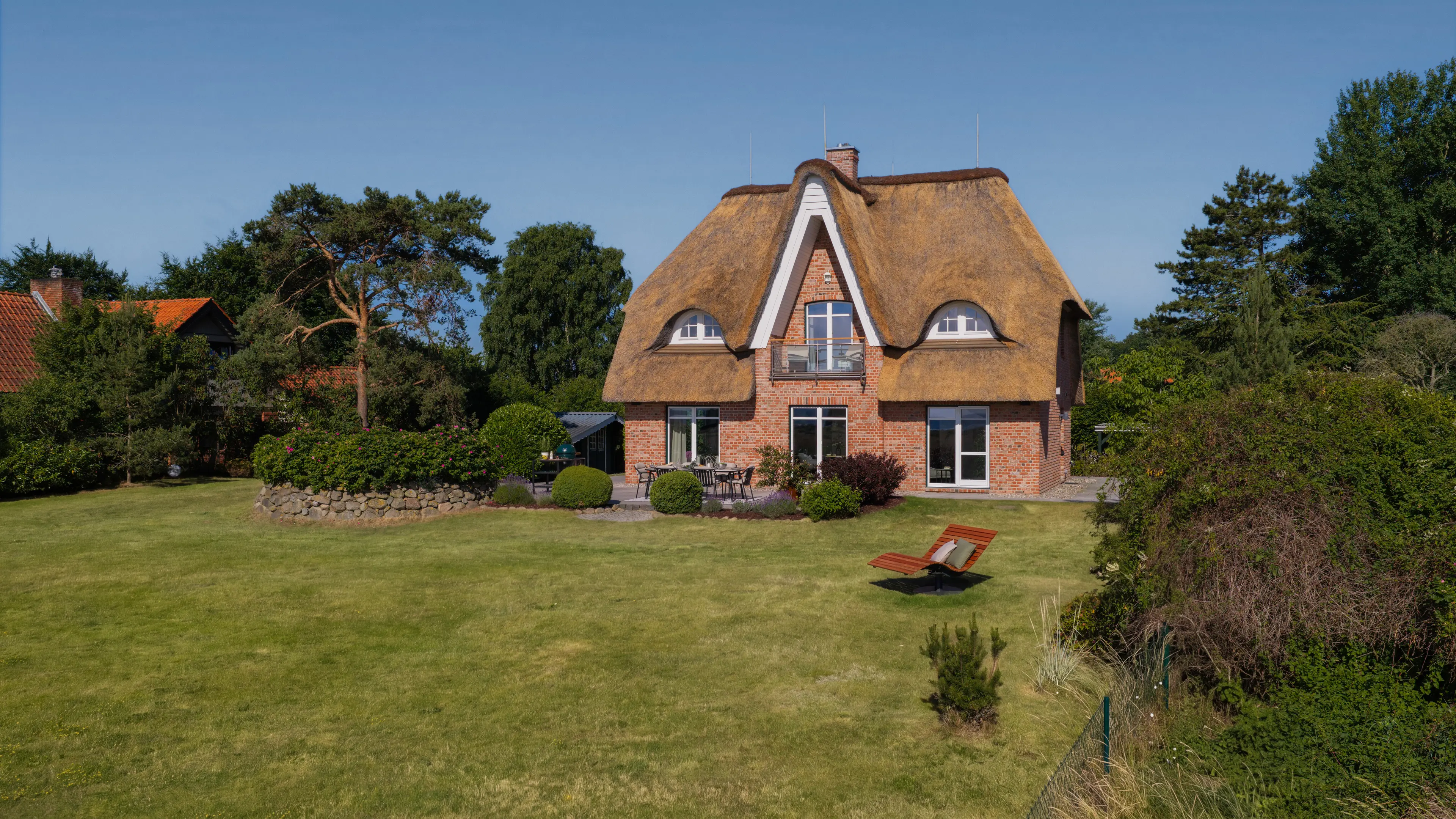 Luftaufnahme des Traumhauses von der Gartenseite: reetgedecktes Backsteinhaus mit weitläufigem Rasen und einer Holzliege im Vordergrund.