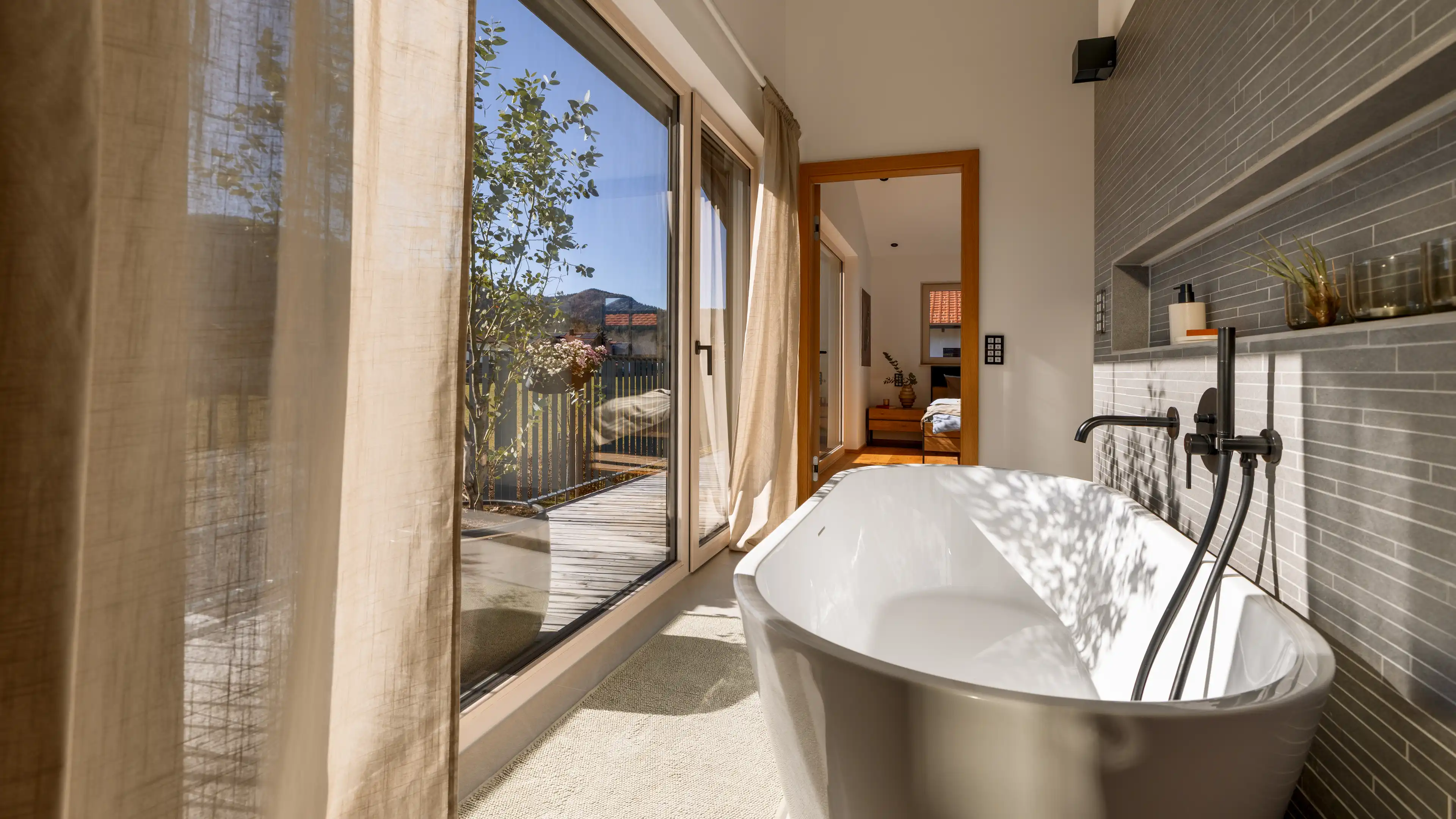 Die große Fensterfront auf der linken Seite des Bildes wirft strahlendes Sonnenlicht ins Badezimmer im Traumhaus am Chiemsee. Direkt am Fenster, mit Blick auf die Terrasse, steht eine weiße freistehende Badewanne vor einer grau gefliesten Wand mit eingebauten Ablagemöglichkeiten und matt-schwarzer Armatur.