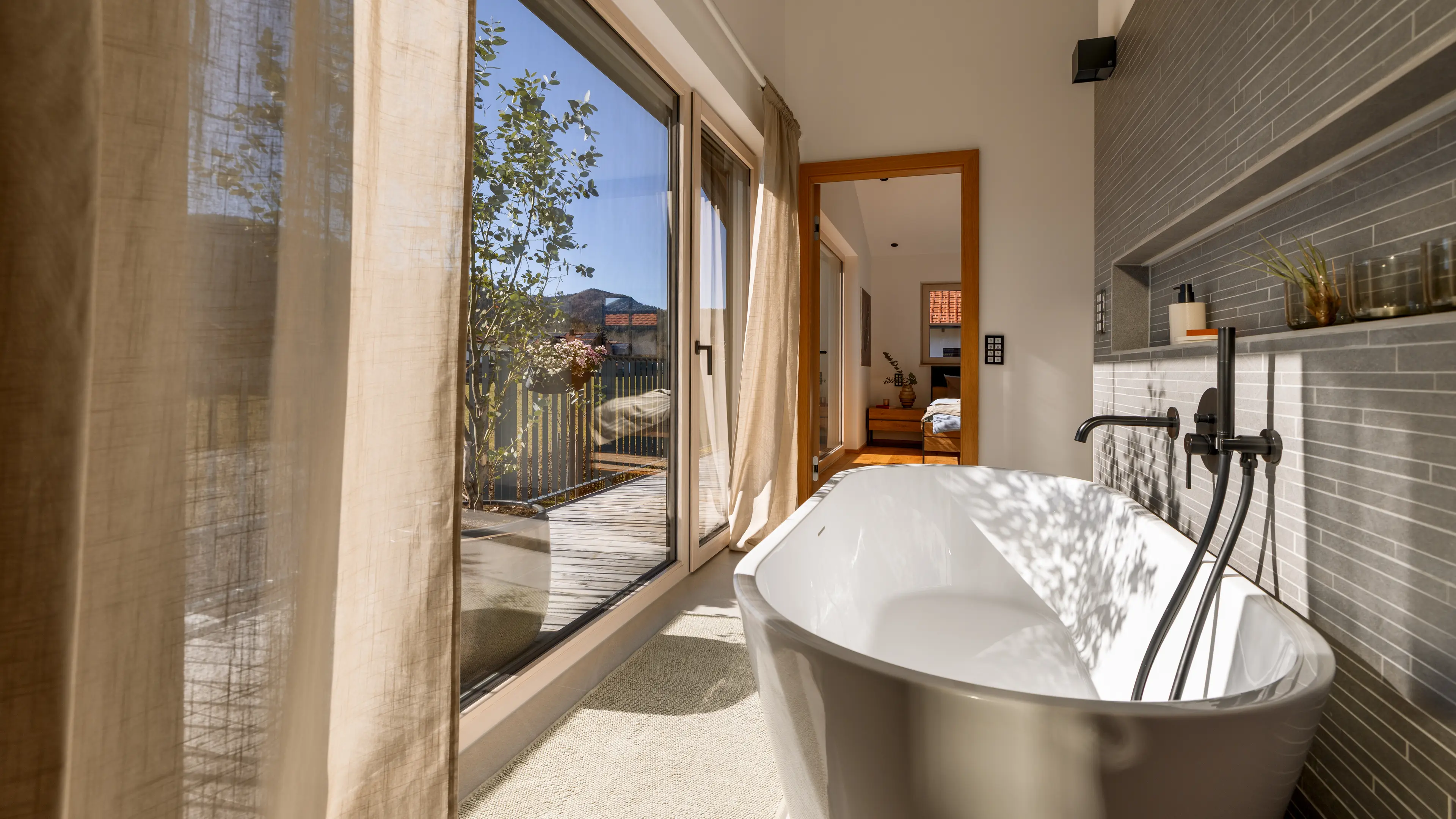 Die große Fensterfront auf der linken Seite des Bildes wirft strahlendes Sonnenlicht ins Badezimmer im Traumhaus am Chiemsee. Direkt am Fenster, mit Blick auf die Terrasse, steht eine weiße freistehende Badewanne vor einer grau gefliesten Wand mit eingebauten Ablagemöglichkeiten und matt-schwarzer Armatur.