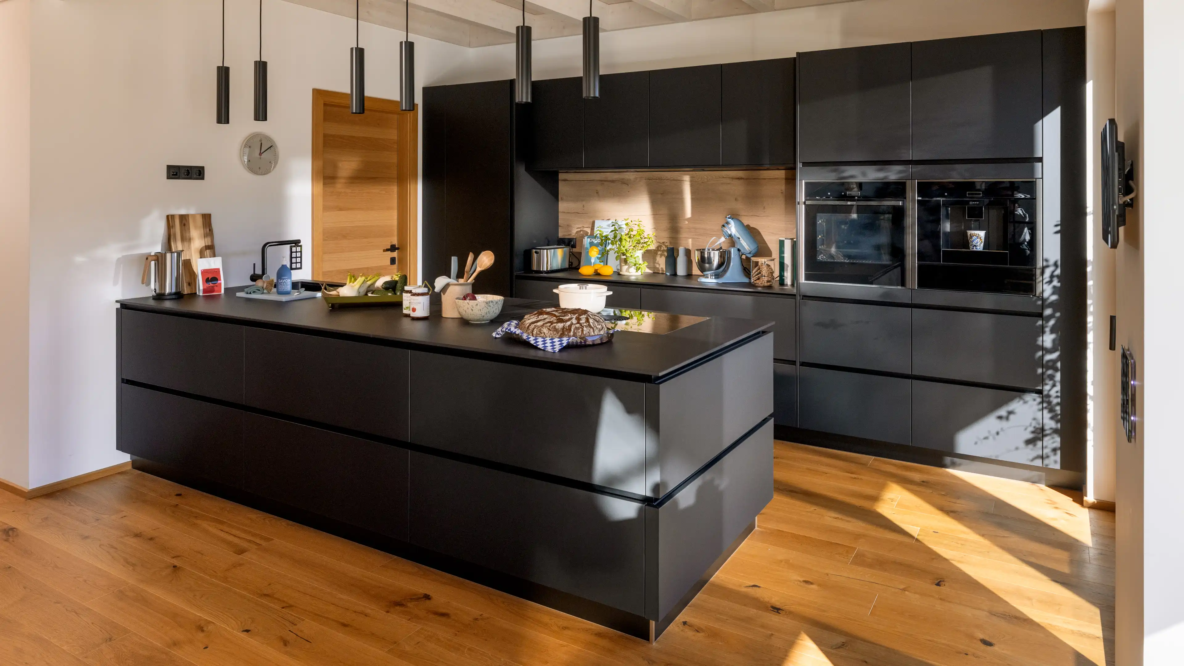 Blick auf die moderne schwarze Küche mit großer Kochinsel und Keramikarbeitsplatte, Bora-Induktionskochfeld und integriertem Kaffeeautomaten im Traumhaus am Chiemsee.