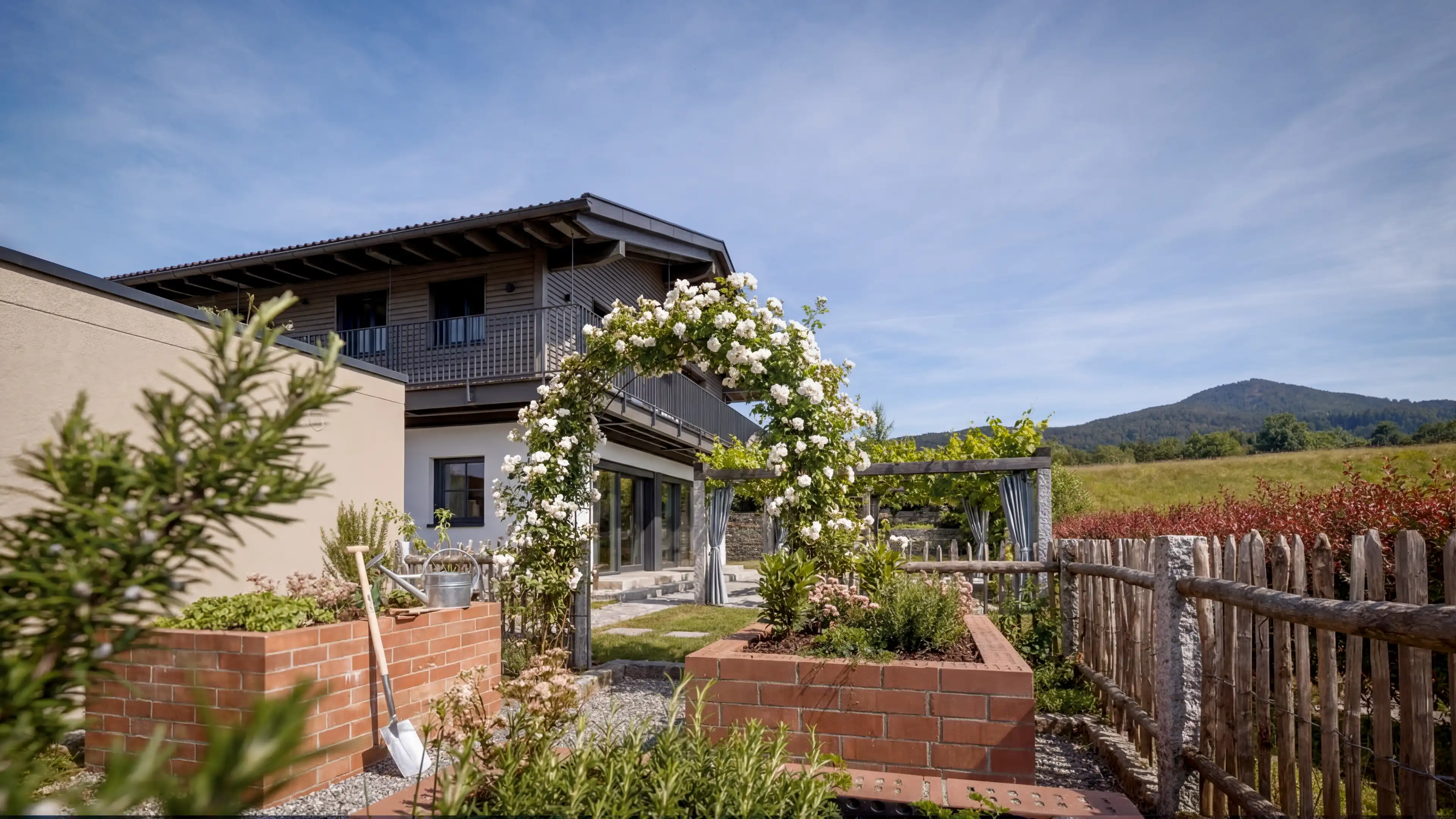 Blick auf die 3 von 4 Klinkerhochbeete des Traumhauses am Chiemsee. Die Hochbeete sind mit unterschiedlichen Kräutern bepflanzt, gegen eines der Beete lehnt eine Schaufel. Der Bauerngarten ist von einem Holzzaun umgeben, der Eingang ist von einem Rosenbogen geschmückt. Im Hintergrund, ist das Traumhaus am Chiemsee und das Bergpanorama zu sehen.