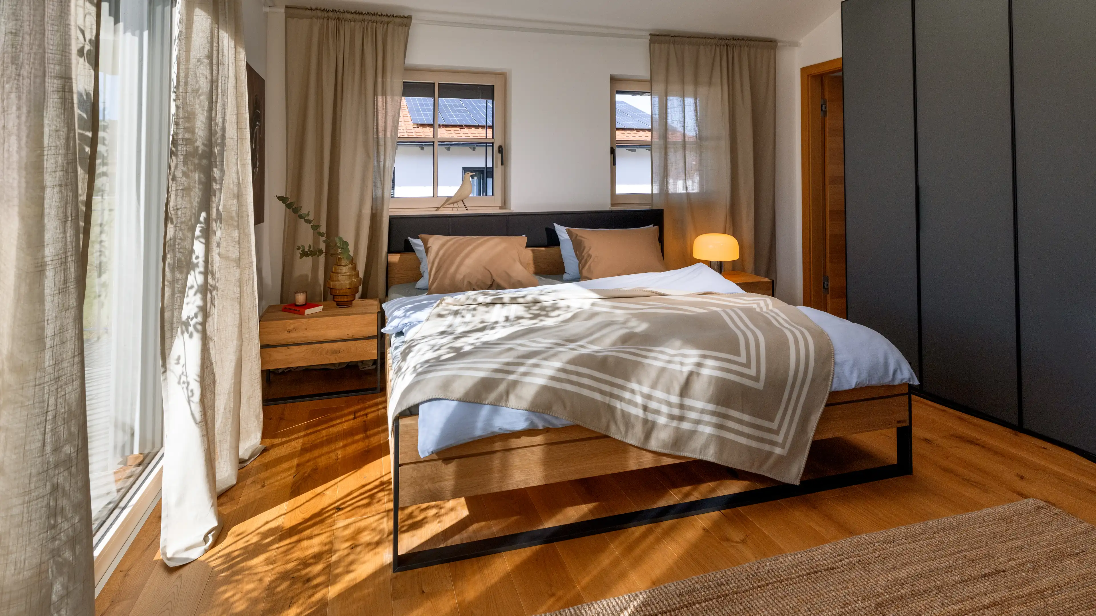 Das Hauptschlafzimmer des Traumhauses am Chiemsee besteht aus einem großen Doppelbett in einer modernen Holzoptik mit schwarzen Metallelementen ist der Mittelpunkt des Bildes. Eine helle Bettwäsche mit beigen Akzenten bringt Gemütlichkeit in den Raum. Hinter dem Bett sind zwei Fenster. Rechts und links vom Bett zwei schwebende Nachschränke aus Holz auf denen ein Buch, eine Zimmerpflanze und eine kleine Tischlampe dekoriert sind.