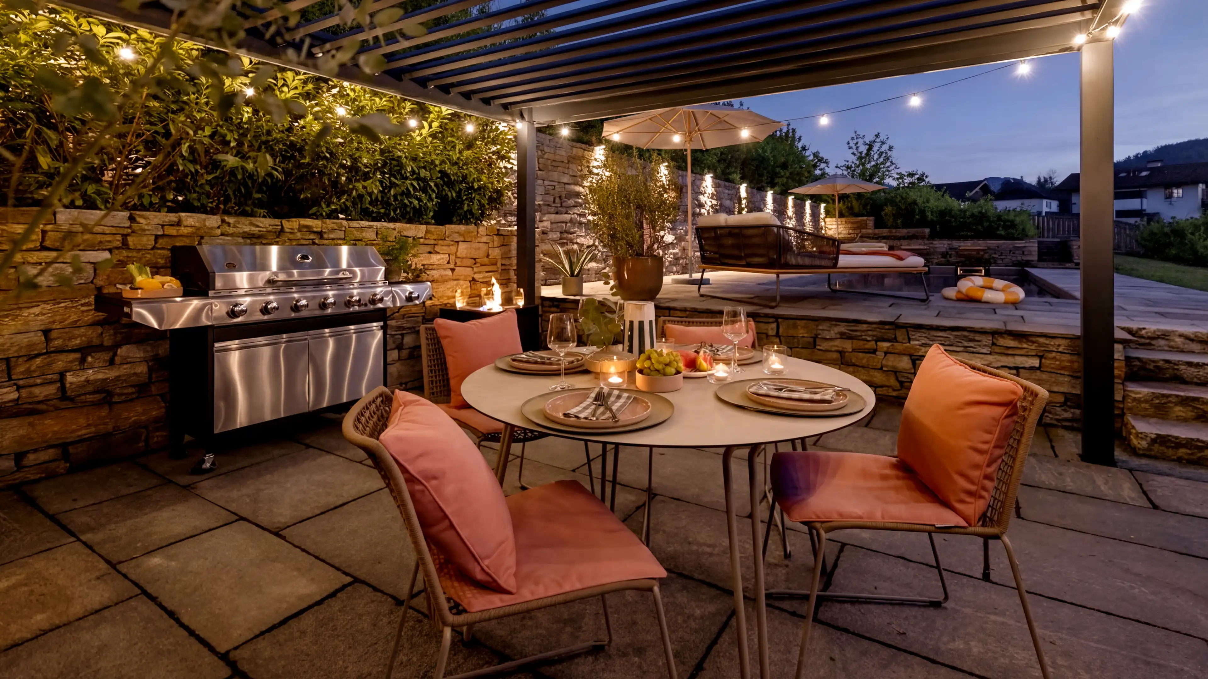 Umgeben von einer Natursteinmauer und mit Blick auf den Pool befindet sich der vertiefte überdachte BBQ- mit Sitz-Bereich des Traumhauses am Chiemsee. Ein heller runder Tisch umgeben von vier Stühlen mit lachsfarbenden Zierkissen ist für einen Grillabend eingedeckt. Auf der rechten Seite hinter dem Tisch steht ein großer silberner Weber Grill. Es wird bereits dunkel und die Poollichter als auch das installierte Licht im BBQ-Bereich sind bereits an.