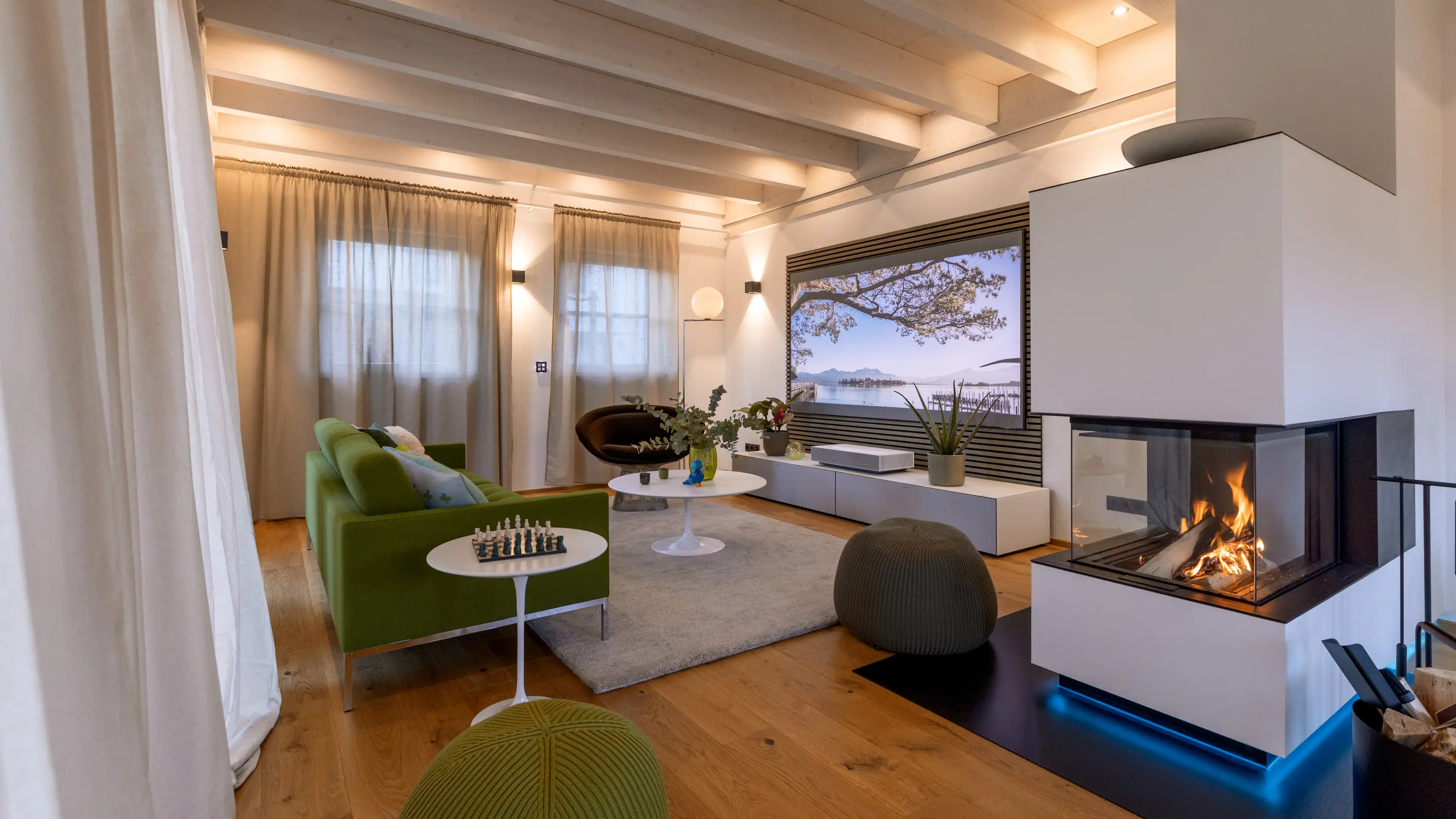 Helles offenes Wohnzimmer im Traumhaus am Chiemsee mit viel indirekter Beleuchtung, grünen Sitzgelegenheiten und weißen Möbelstücken. Vom Sofa geht der Blick direkt auf die große Leinwand und das dazugehörige Surround System. Rechts neben den Sofa öffnet sich der Raum zum Essbereich, als Raumtrenner sorgt der von 3 Seiten verglaste weiße Kamin, in dem ein Feuer lodert.