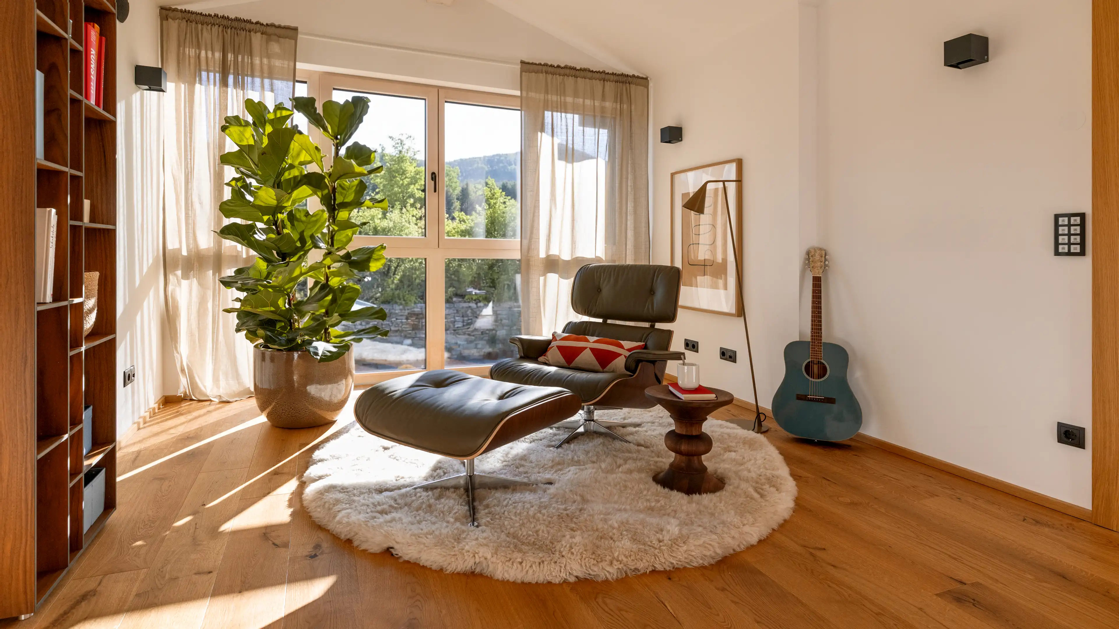 Der schwarze Vitra Lounge Chair ist das Zentrum des Bildes. Dieser steht auf einem hellen, runden und flauschigen Teppich. Neben dem Stuhl steht ein kleiner Beistelltisch auf dem ein Buch und ein Glas Wasser steht. Im Hintergrund bringen die bodentiefen Fenster Licht in den Raum. Eine große grüne Zimmerpflanze, ein beiges abstraktes Bild an der weißen Wand, eine dunkelgrüne Gitarre und ein Stehlampe runden die Gallerie im Traumhaus am Chiemsee ab.