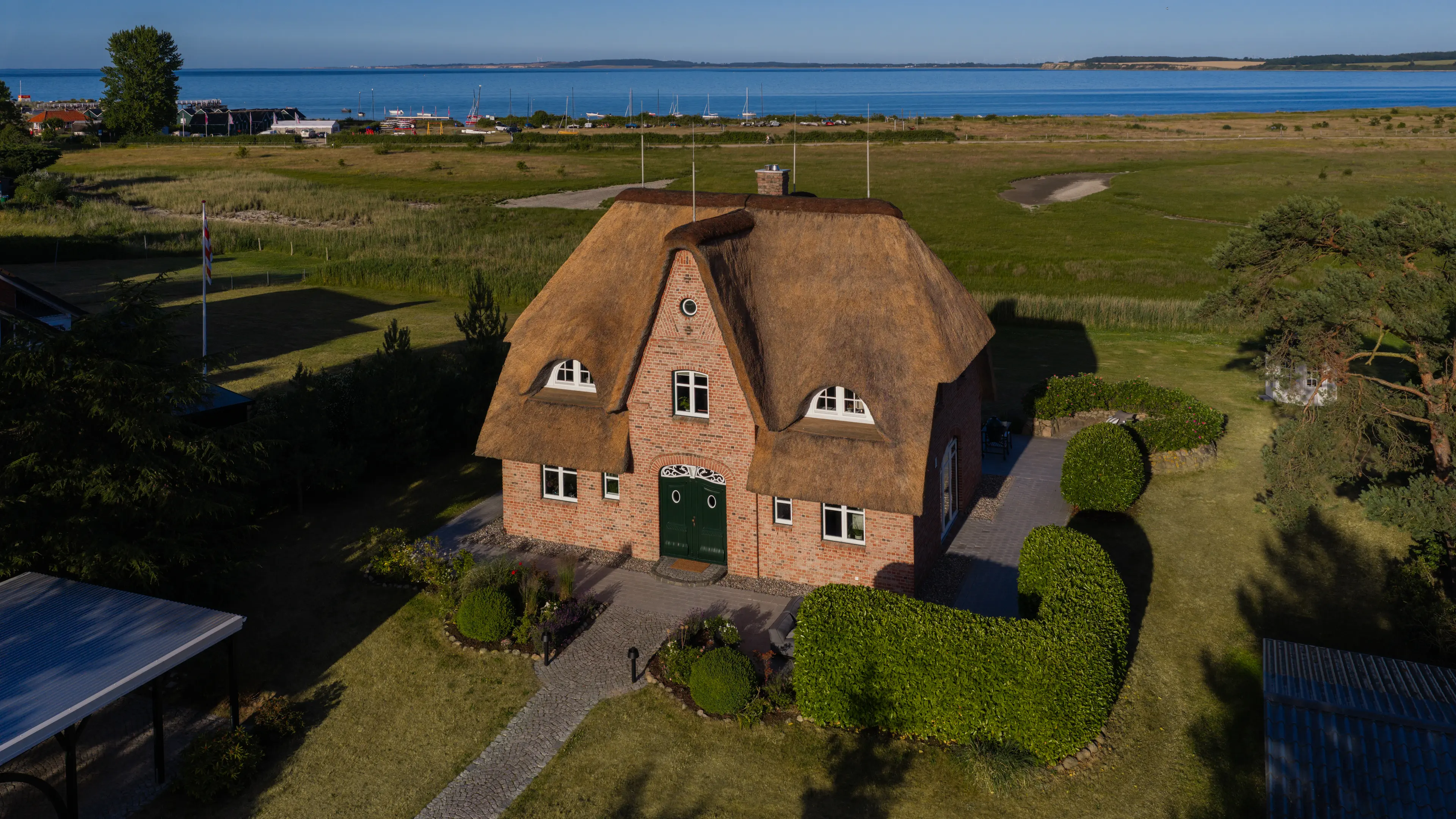 Das Traumhaus mit Ostseeblick aus der Vogelperspektive seitlich mit Blick auf das rote Backsteinhaus, weißen Fenstern, dunkelgrüner Eingangstür und Reetdach. Um das Traumhaus herum erstreckt sich ein großer grüner Garten mit einigen Büschen und viel Rasenfläche. Hinter dem Grundstück ist ein Naturschutzgebiet und die Ostsee zu sehen.