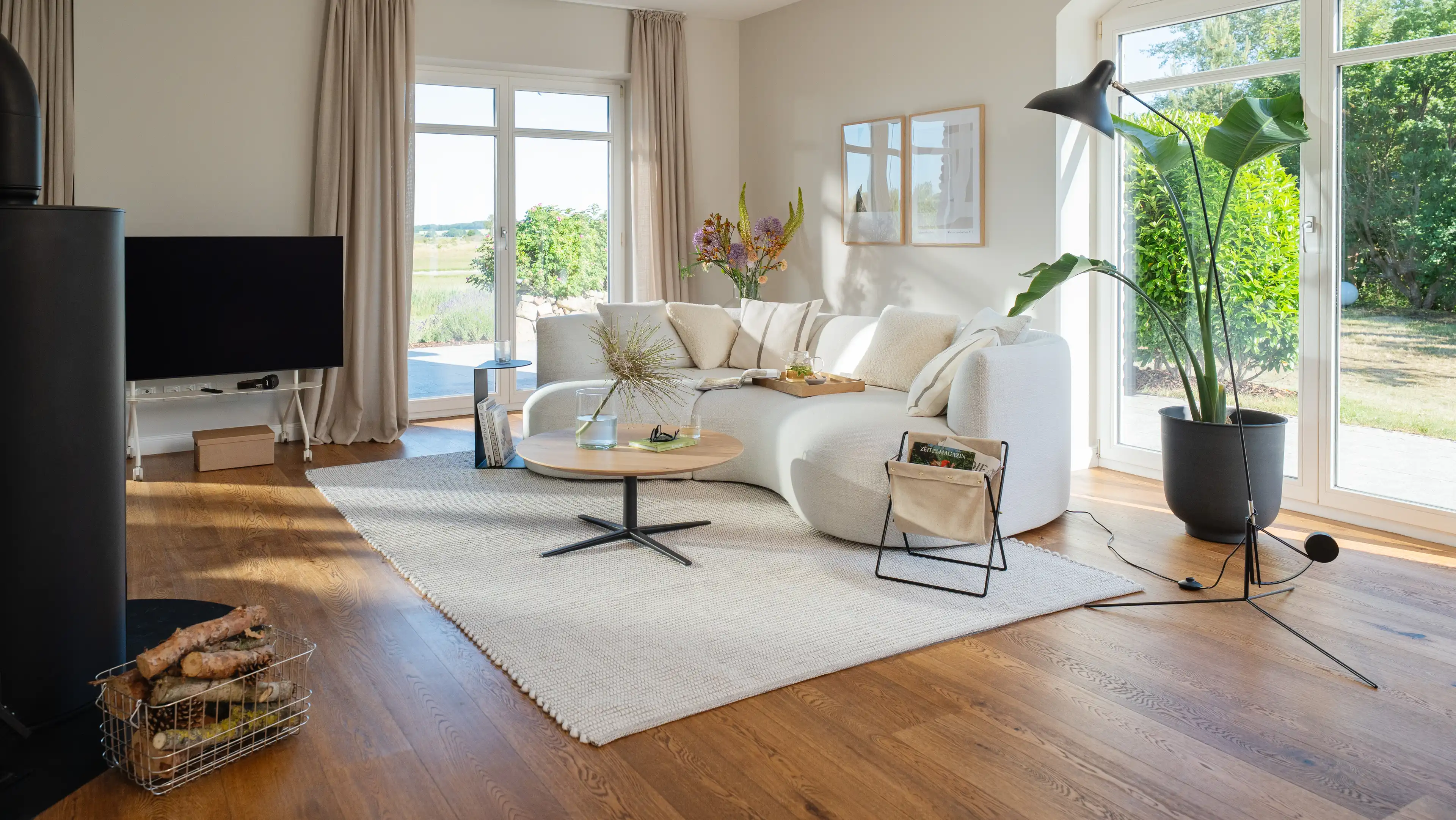Wohnbereich mit weißem geschwungenem Sofa, rundem Holztisch, Kaminofen (Feuer hier nicht sichtbar entzündet) und Korb mit Brennholz.