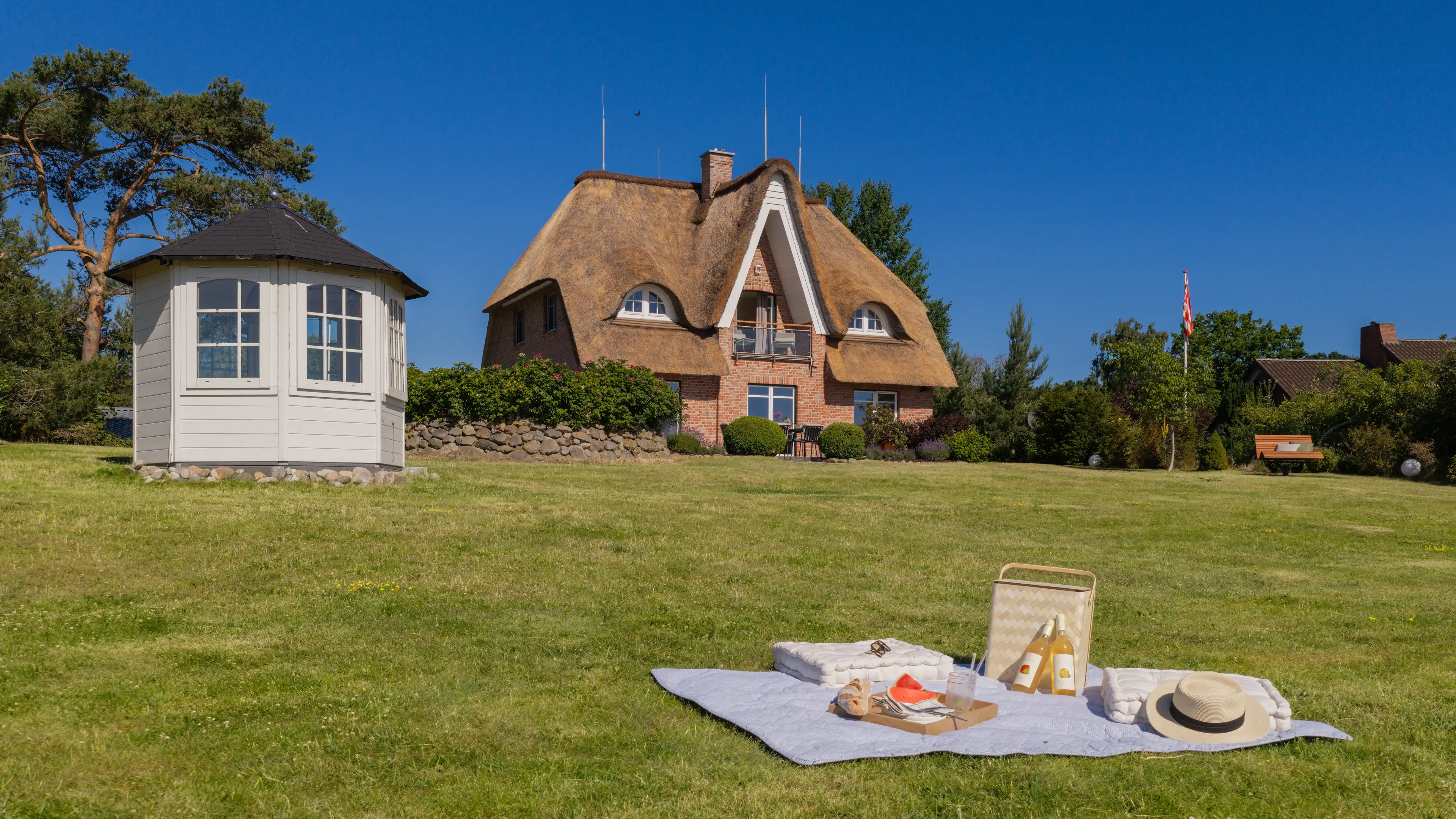 Weitläufiger Garten mit weißem Gartenpavillon, gedecktem Picknick auf einer Decke im Vordergrund und dem Traumhaus im Hintergrund.