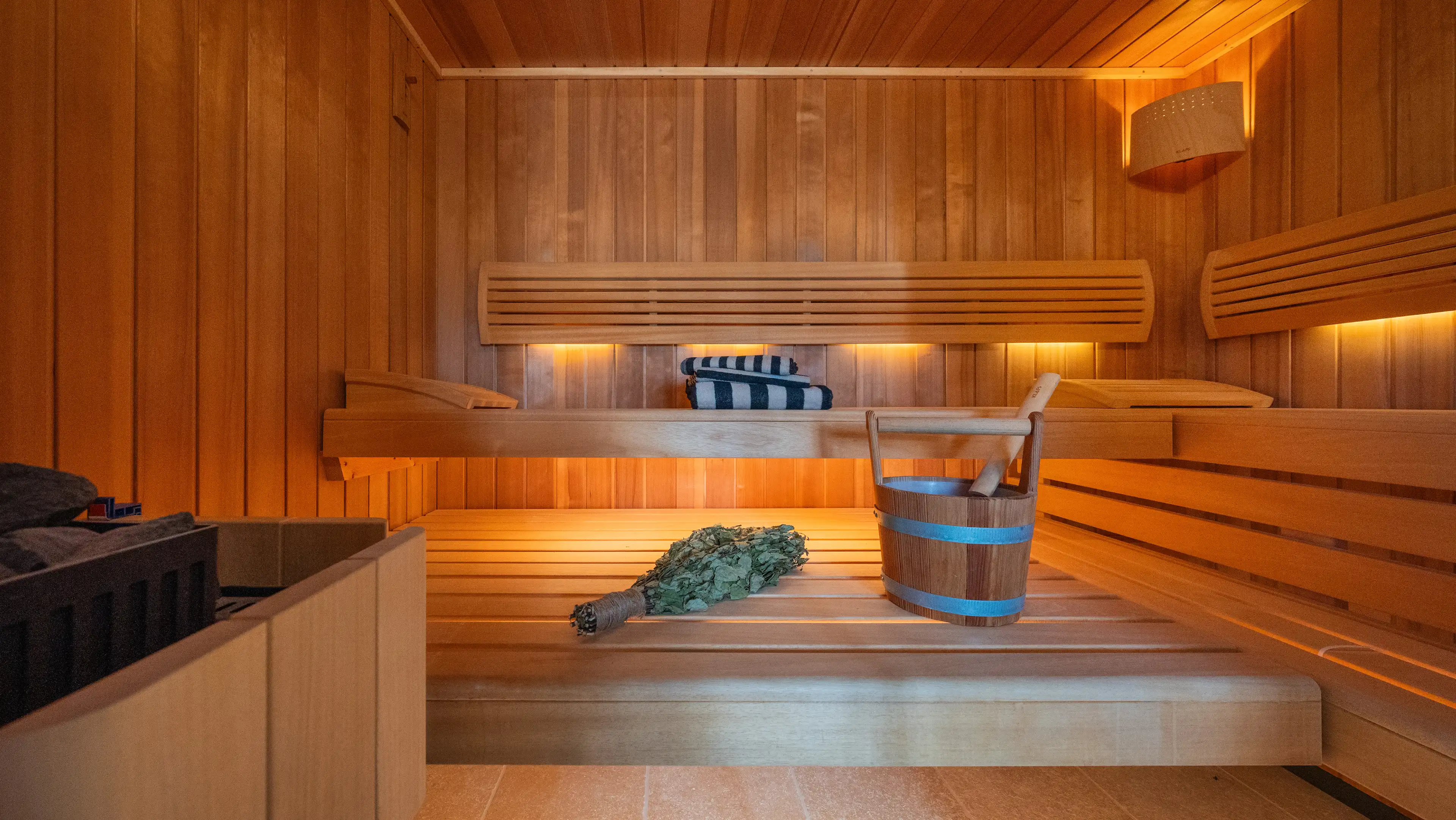 Holzverkleidete Sauna mit Bänken, gerollten Handtüchern, einem Holzkübel mit Aufgusskelle und einem Birkenzweig-Bündel.