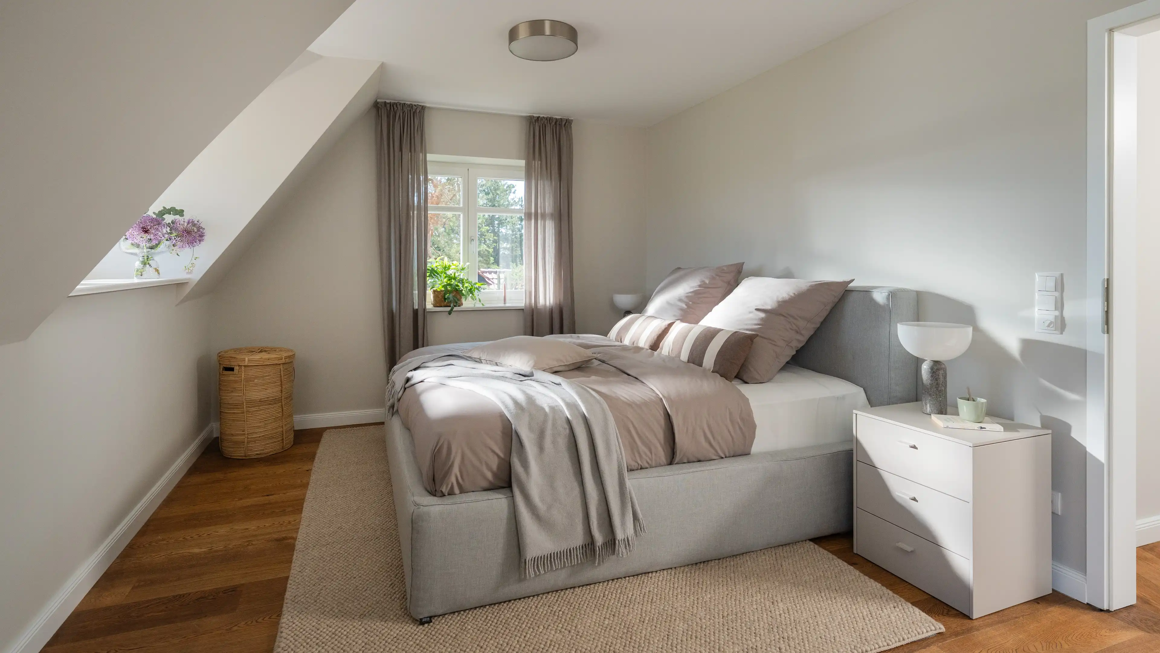 Helles Schlafzimmer unter Dachschräge mit gepolstertem grauem Bett, taupefarbener Bettwäsche, weißem Nachttisch und einem Rattankorb.