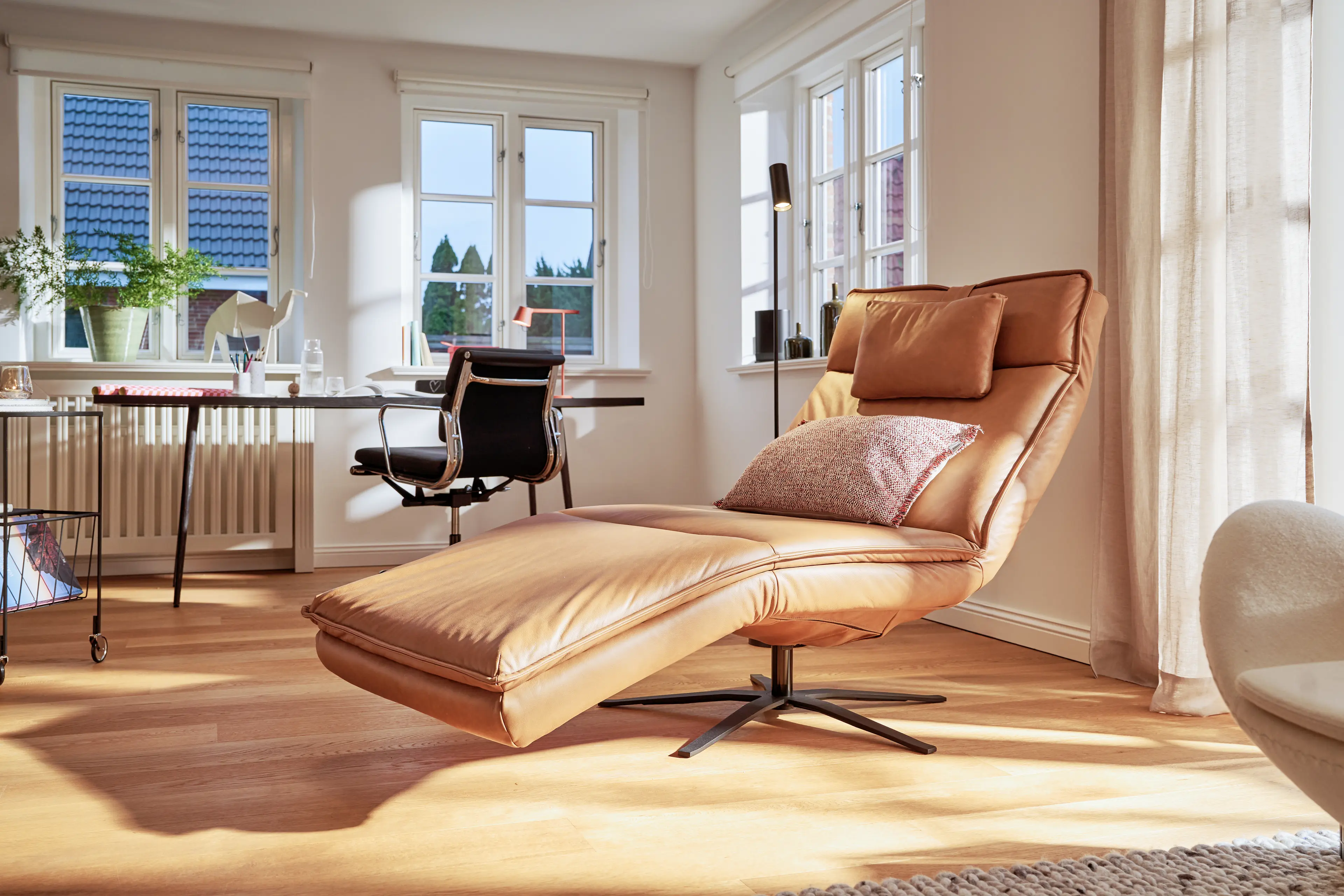 Arbeitsplatz im Wohnzimmer des Traumhauses umgeben von Tageslicht mit Schreibtisch und schwarzen Lederstuhl. Im Vordergrund befindet sich außerdem ein mocca-farbende Relaxliege Belize von SWK.