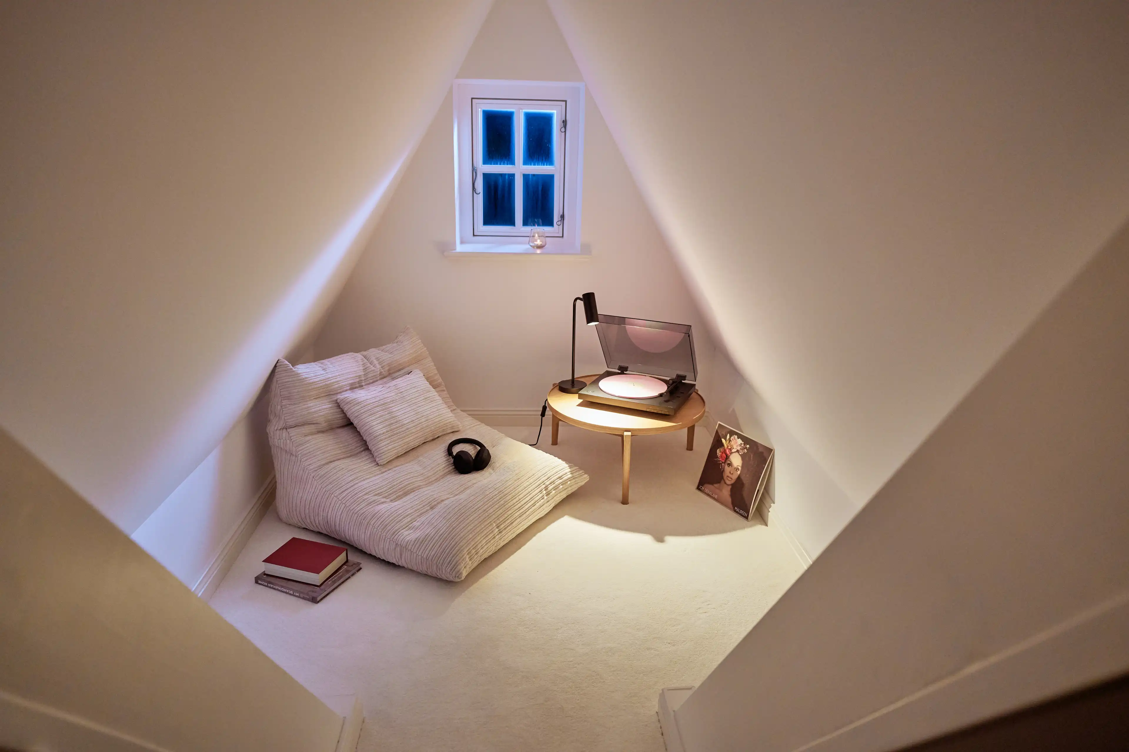 Ausgebauter Spitzboden mit kleinem Fenster, ganz in weiß gehalten mit gemütlicher moderner Sitzgelegenheit, Plattenspieler und Büchern.