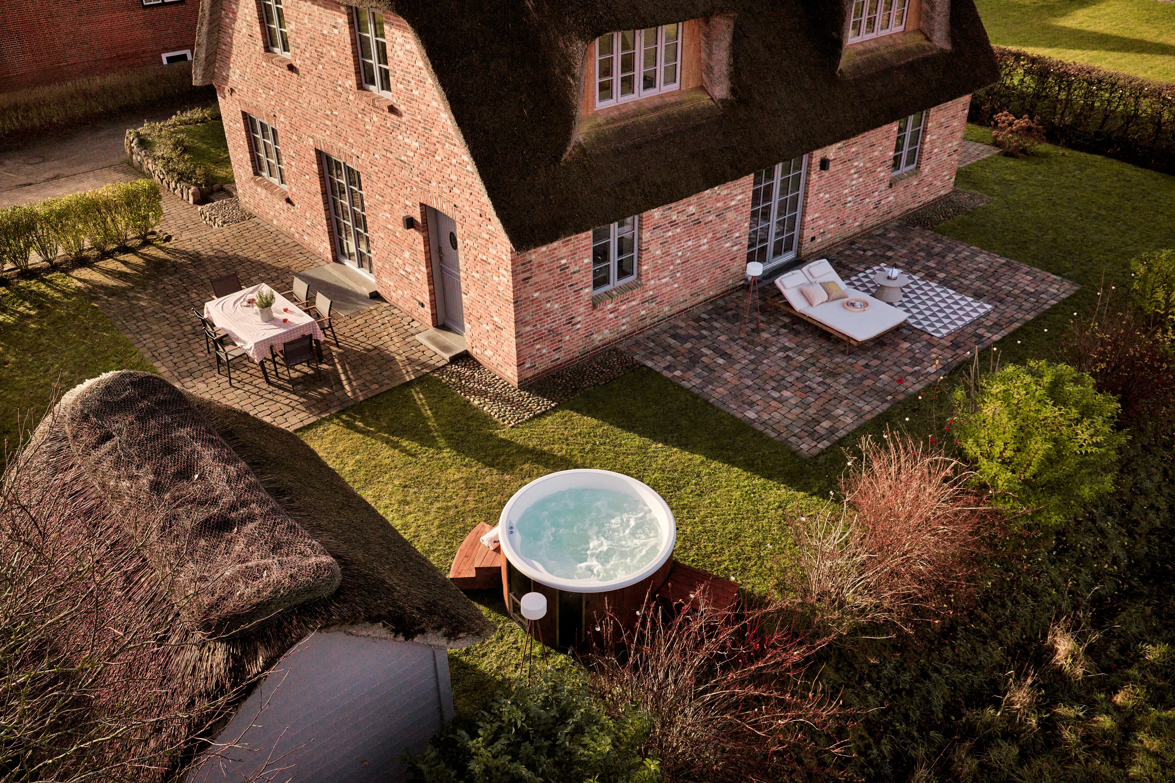 Vogelperspektive auf den Garten des Traumhauses auf Föhr mit Blick auf 2 der 3 Terrassen inkl. Gartenhäuschen, gedeckter Esstisch, Hot Tub, Sonnenliegen und grüne Rasenfläche.