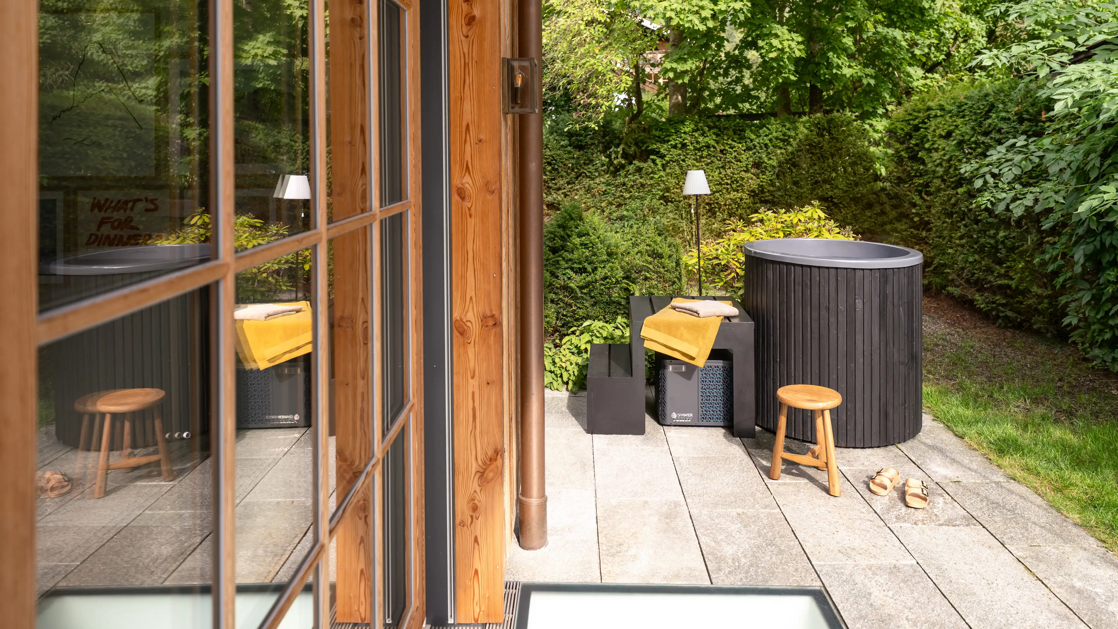 Rundes, schwarz verkleidetes Cold-/Hot-Tub auf einer Steinterrasse neben dem Technikgeraet, mit gelben Handtuechern, einem Holzhocker und dichtem Gruen im Hintergrund.