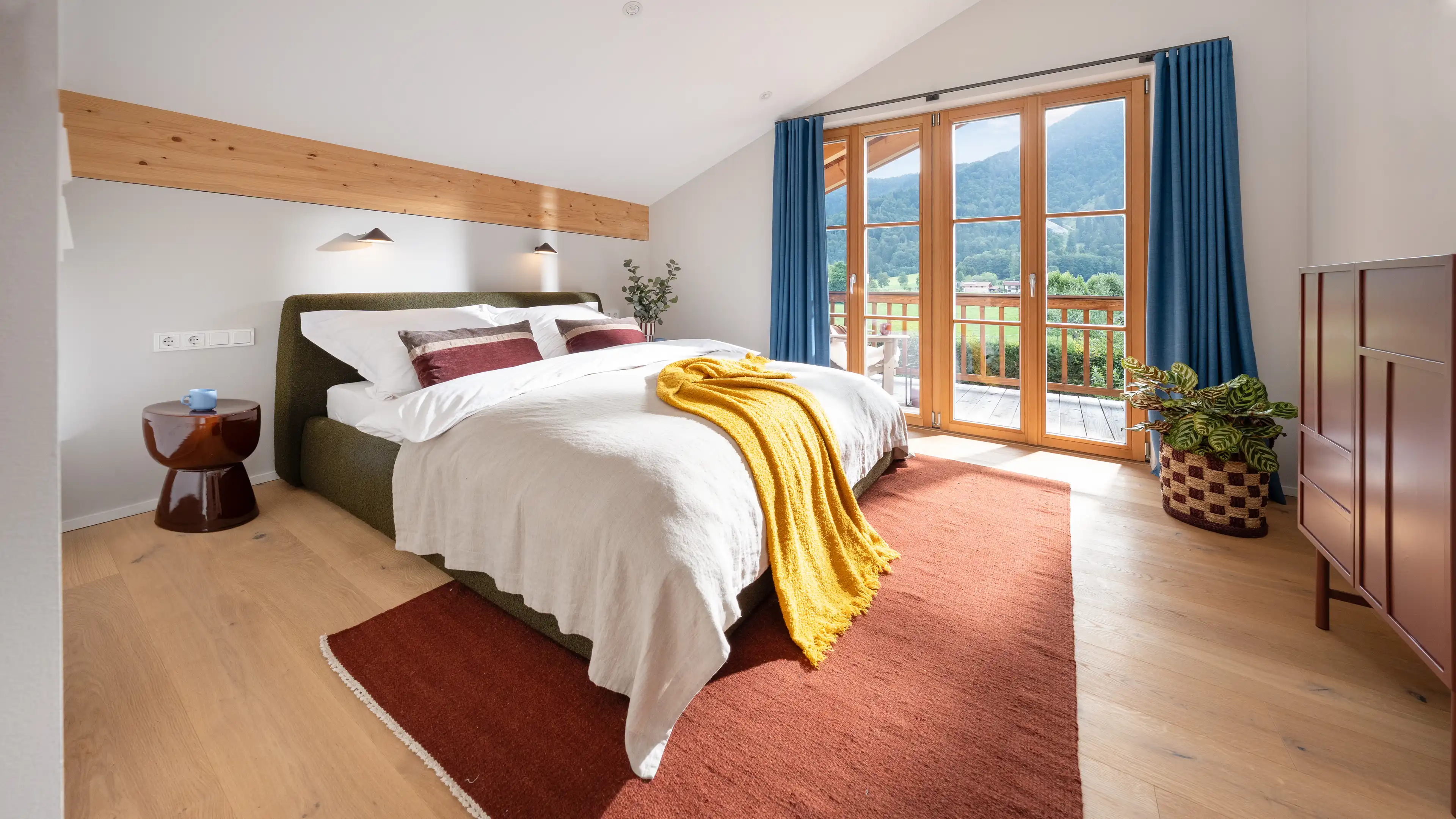 Schlafzimmer mit gruen gepolstertem Bett, weisser Bettwaesche, senfgelber Decke und burgunderroten Kissen, blauen Vorhaengen und Blick durch Glastueren auf Balkon und Berge.