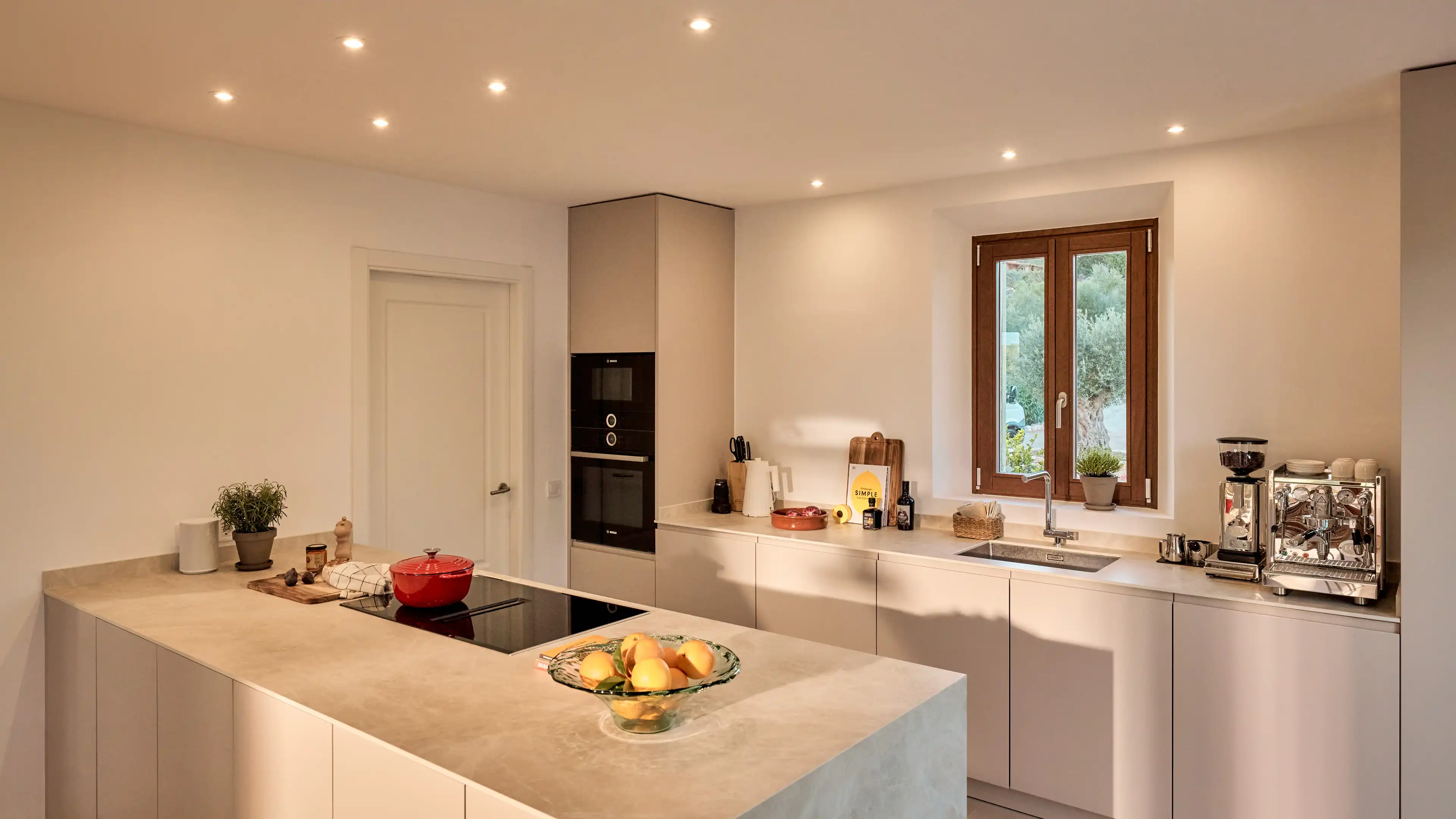Moderne Küche im Traumhaus auf Mallorca mit weißen Fronten und hellgrauer Arbeitsplatte, ausgestattet mit einem Espressomaschine, Ofen auf Augenhöhe und vieles mehr. Von der Küche hat man über ein Fenster direkten Blick in den Garten.