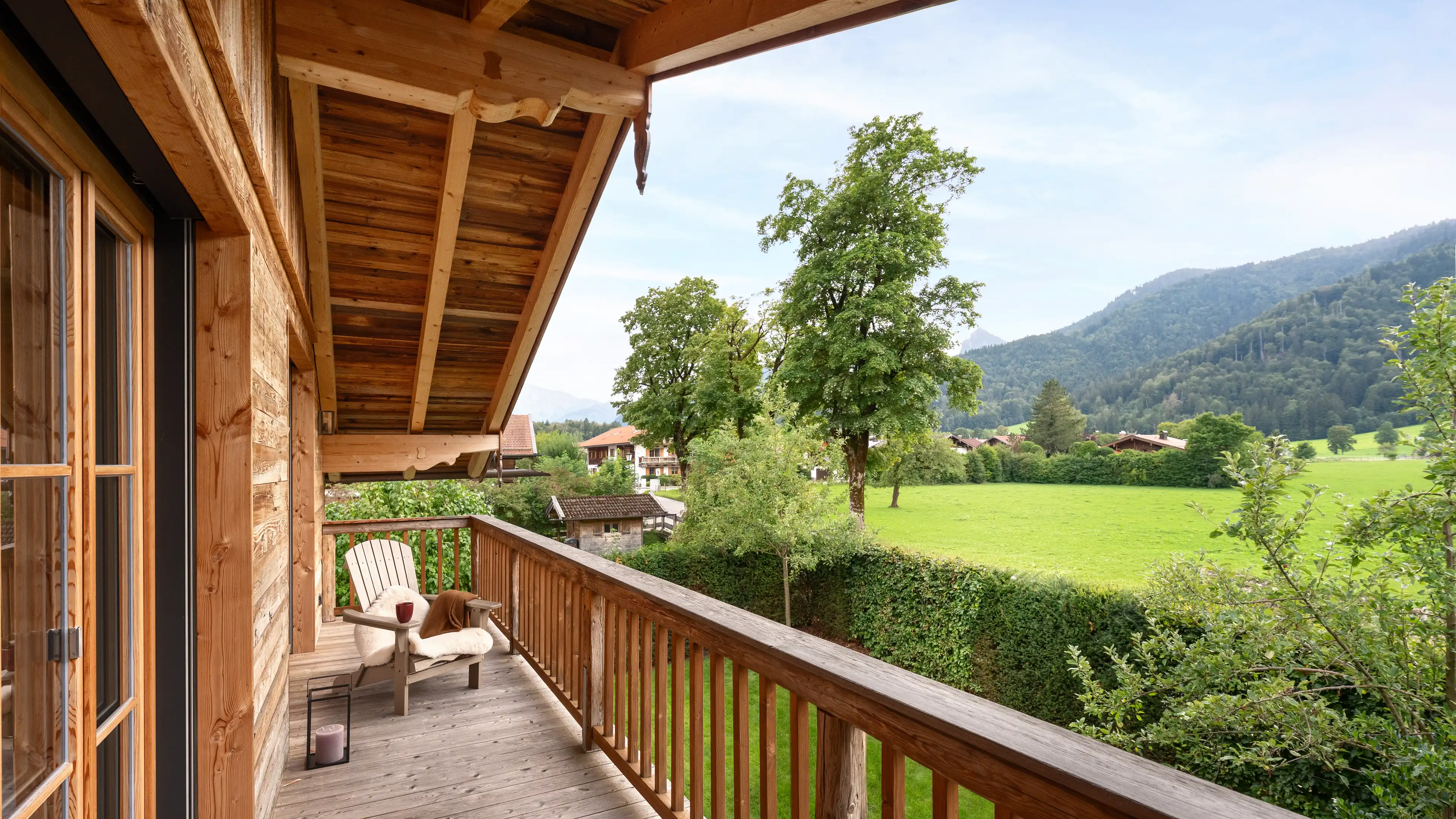 Holzbalkon mit Adirondack-Stuhl, Fellauflage und Laterne, Blick ueber gruene Wiesen und Baeume auf die umliegenden Berge unter blauem Himmel.