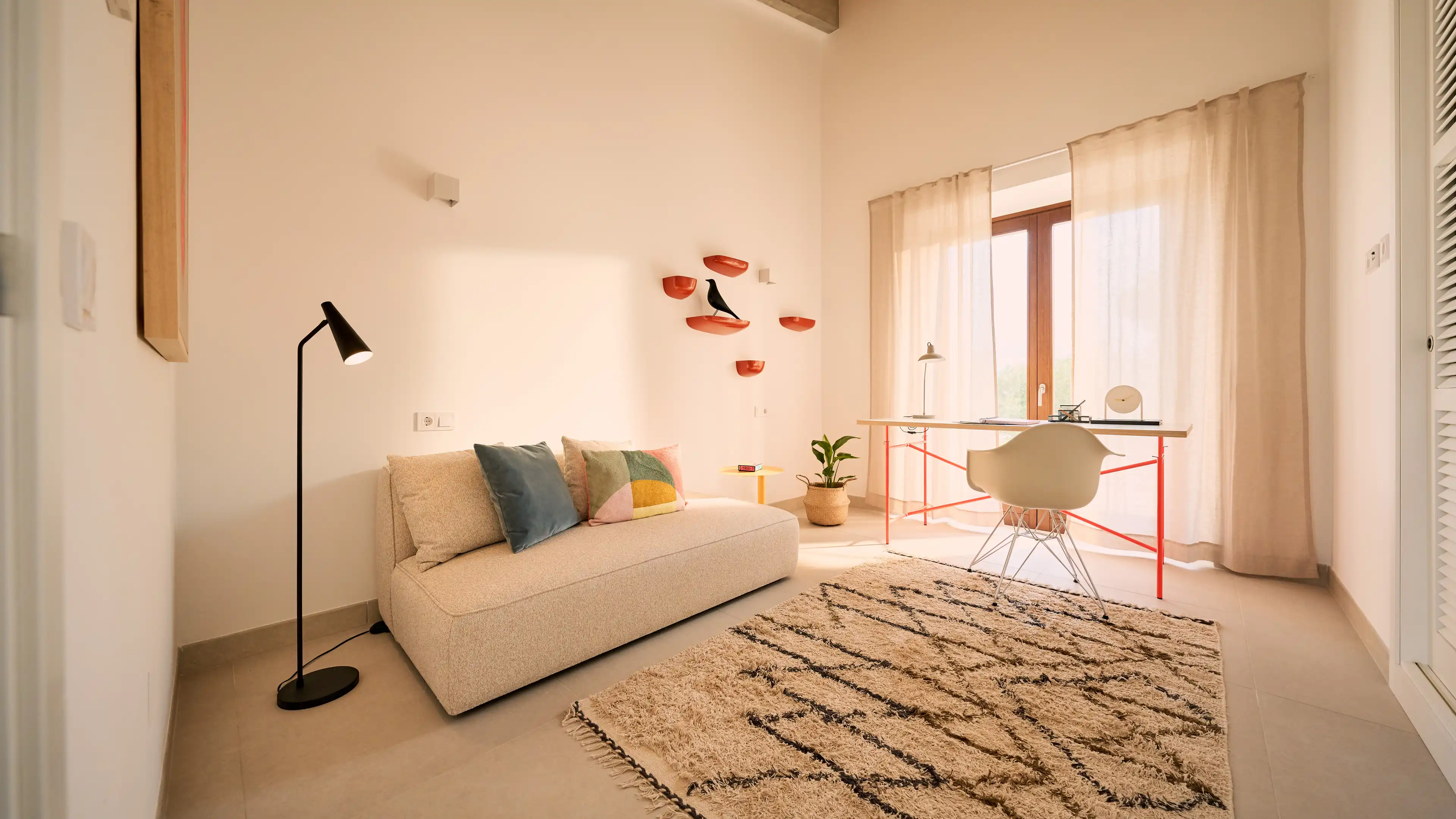Helles Arbeitszimmer im Traumhaus auf Mallorca mit Farbakzenten, weißen Einbauschränken, Schreibtisch direkt vor den bodentiefen Fenstern und einem beigefarbenden kleinen Sofa.