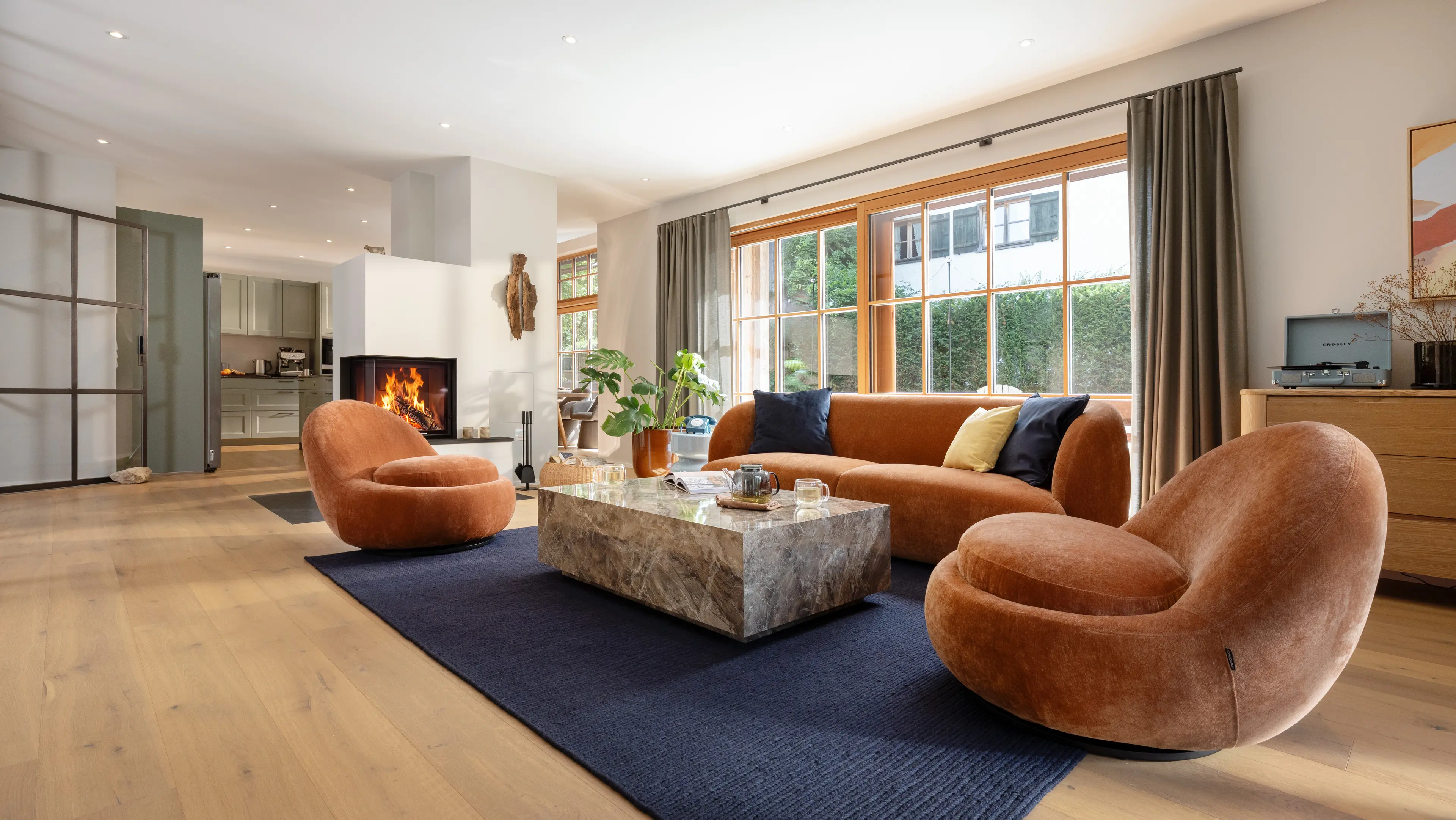 Wohnbereich mit rostorangenem Sofa und zwei runden Sesseln um einen Marmor-Couchtisch, dunkelblauem Teppich und brennendem Kamin vor grossen holzgerahmten Fenstern.