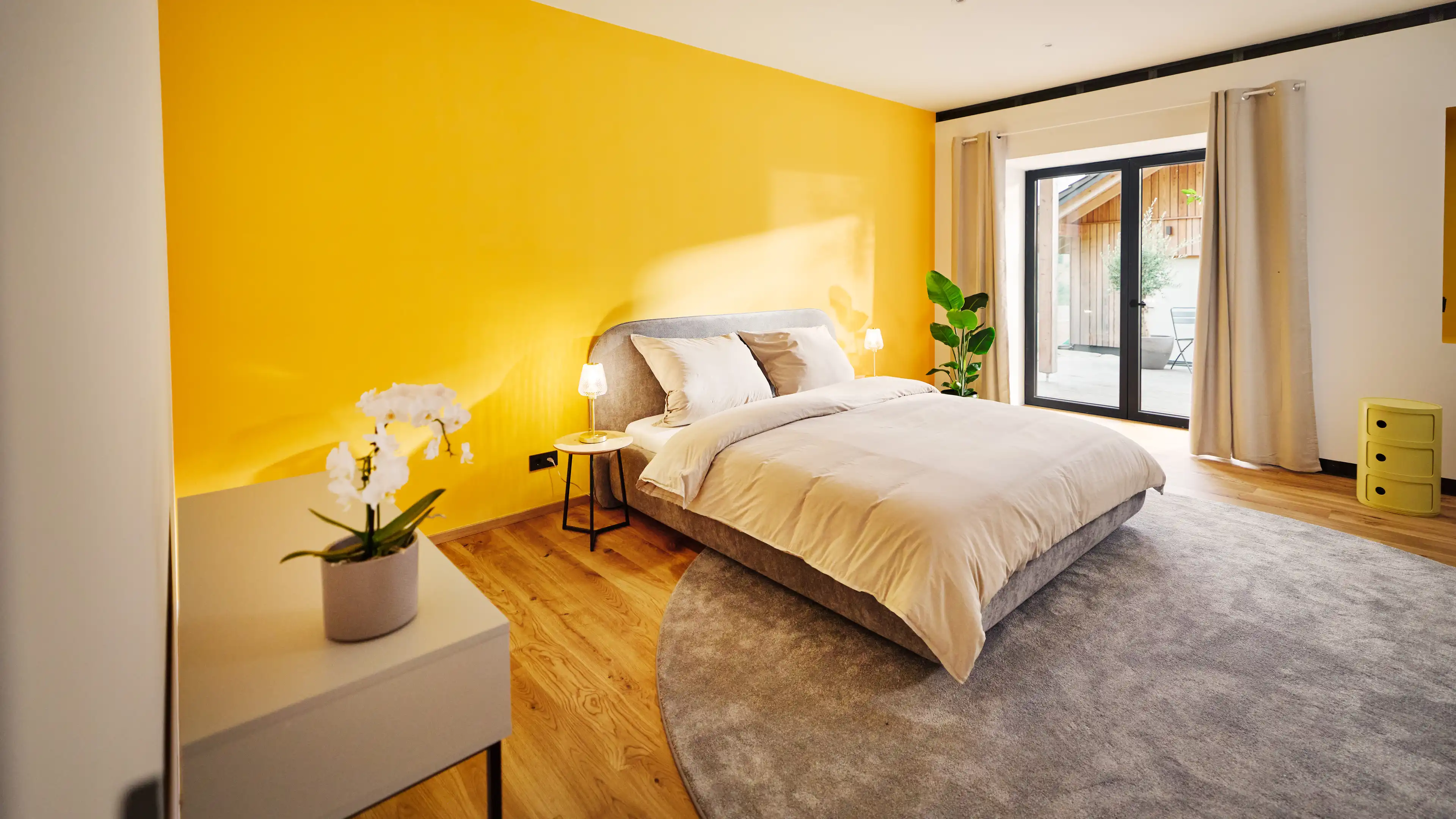 Helles großes Schlafzimmer mit Zugang direkt auf die Terrasse, großes Bett in Grautönen gehalten vor einer sonnengelben Wand