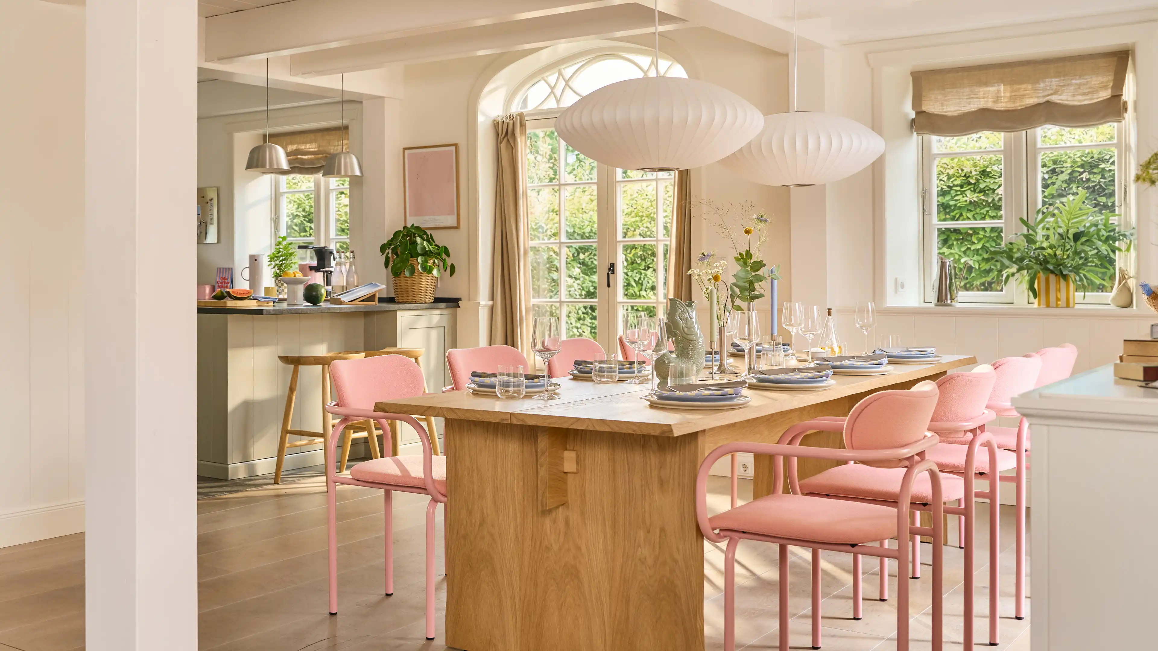 Großer sehr heller Essbereich mit einem hellen langem Holztisch. Am Tisch stehen 8 rosa-farbende Stühle. Im Hintergrund, des Tisches ist ein Teil der offenen weißen Landhausküche zu sehen.