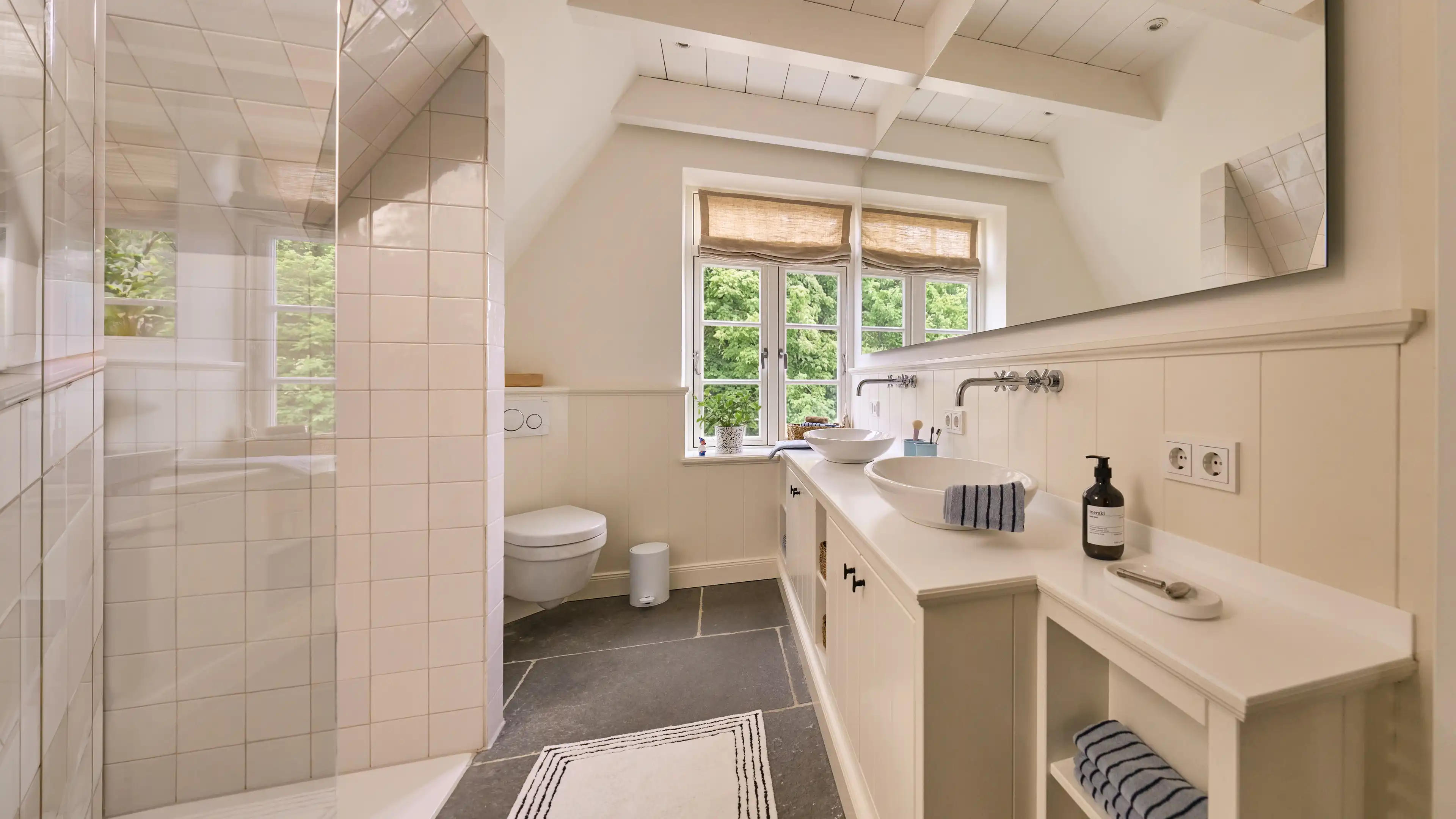 Ein großes Badezimmer mit dunkelgrauen Bodenfliesen und weißen Wandfliesen, 2 große Fenster, 2 Waschbecken auf der rechten Seite und einer verglasten Dusche auf der linken Seite.