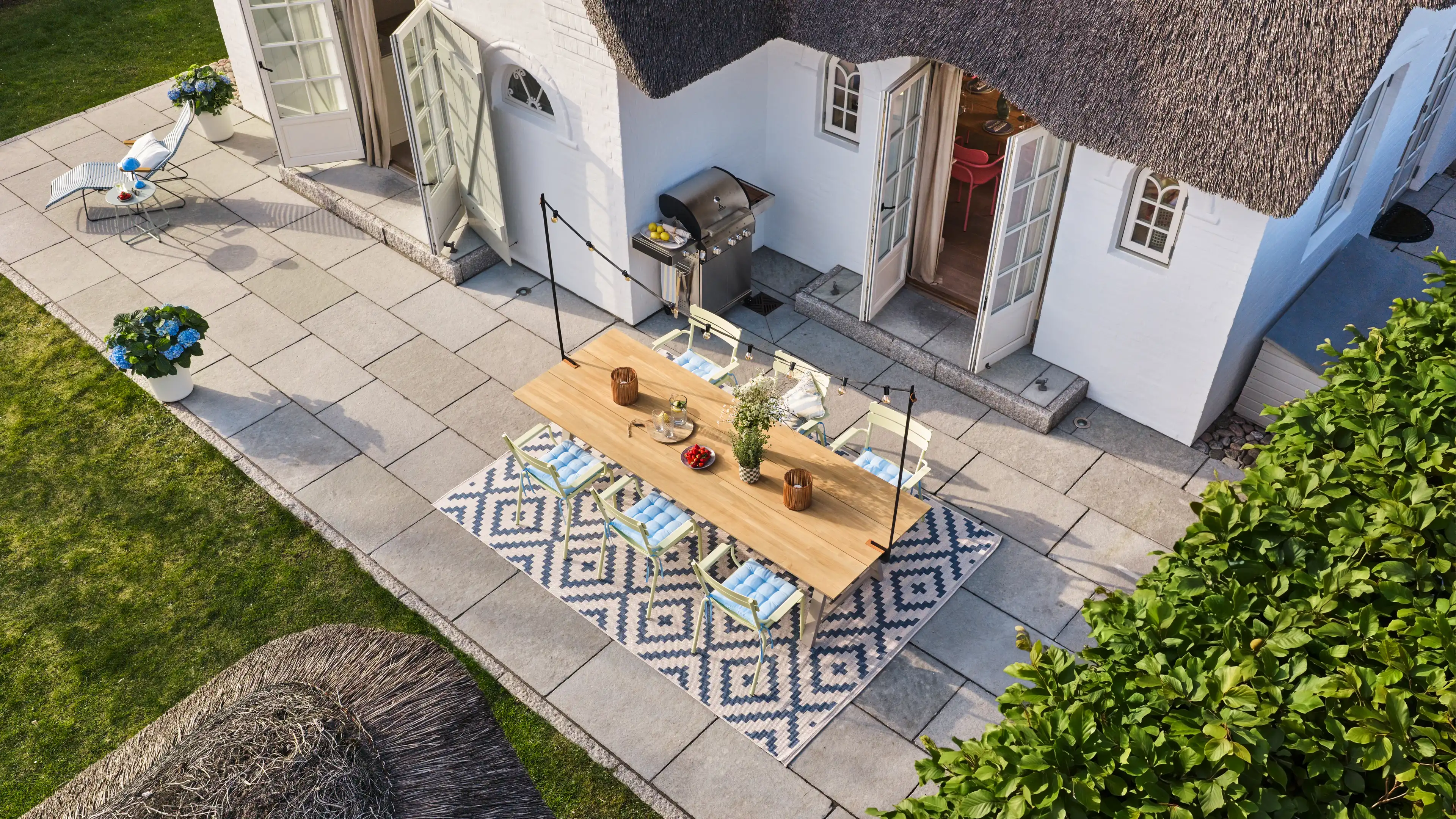 Vogelsperspektive auf die hellgeflieste Terrasse des Traumhauses auf Amrum, dekoriert mit einem Sonnenstuhl und einem großem Holztisch, 6 Stühlen, Outdoor-Teppich und Gas-Grill
