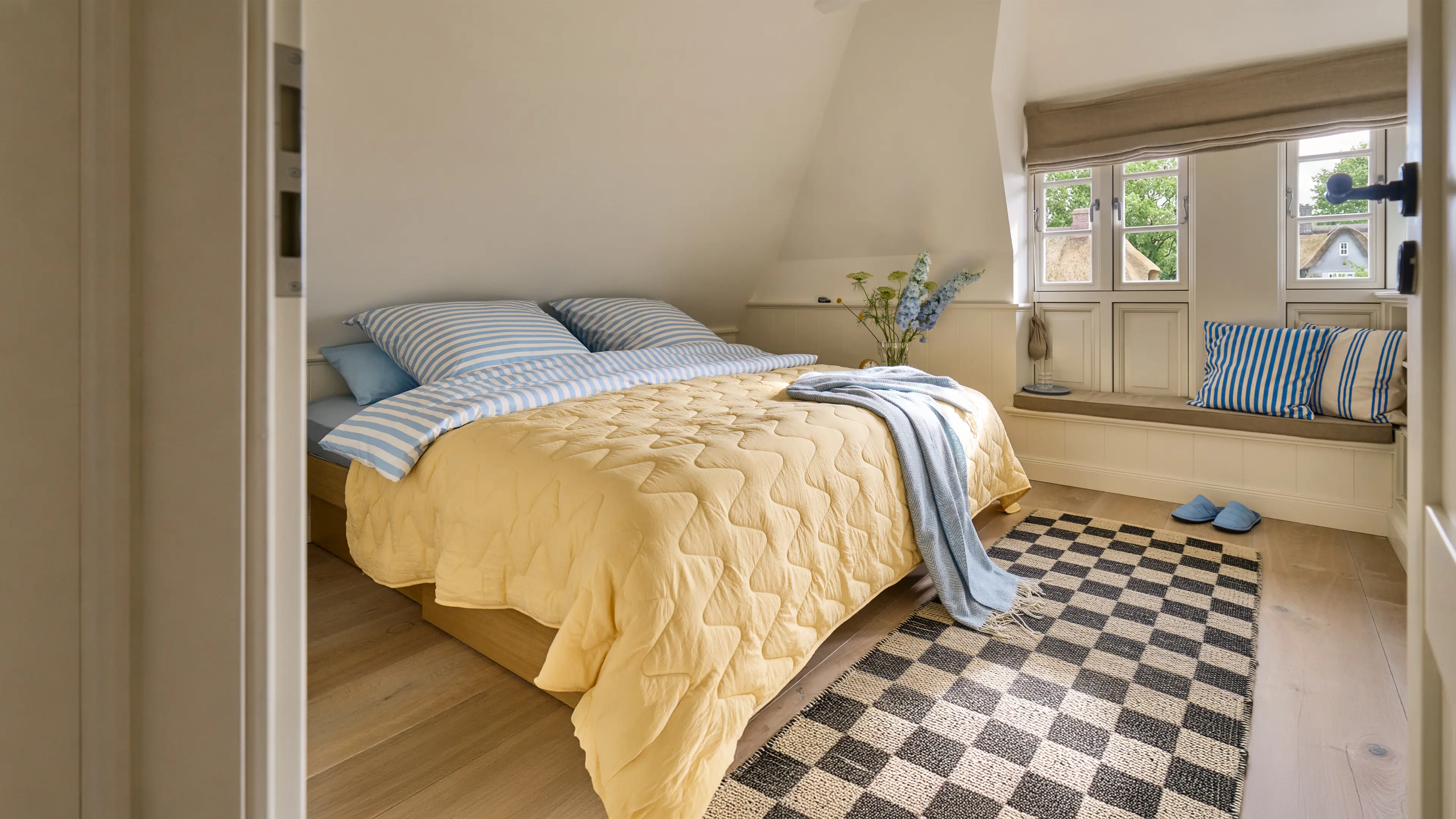 Das Hauptschlafzimmer mit goßem King-Size Bett in pastell blau und gelb gehaltenden Akzenten. Neben dem Bett, ist eine kleine Sitzniche direkt vor den Fenstern zu sehen.