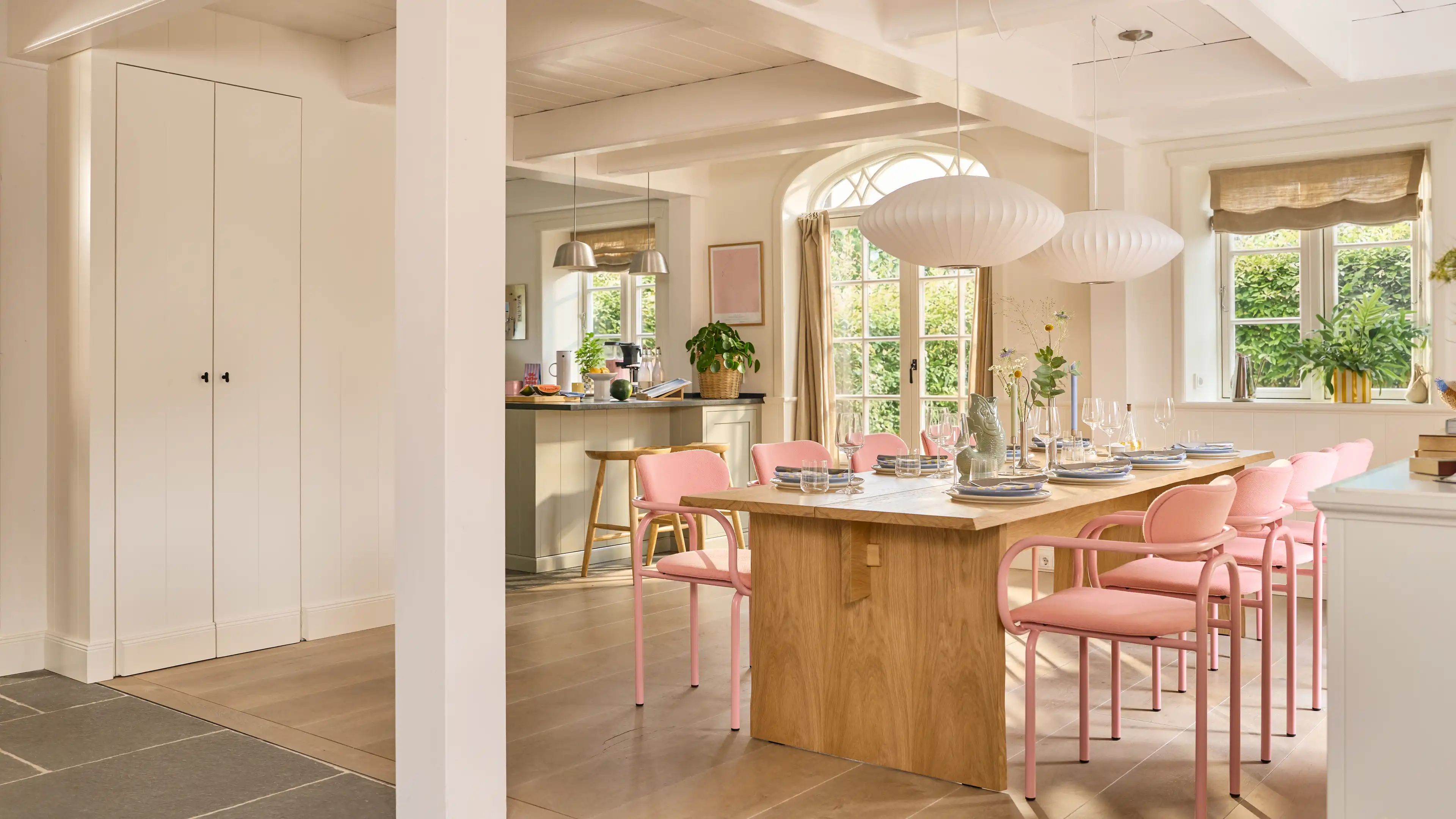 Großer sehr heller Essbereich mit einem hellen langem Holztisch. Am Tisch stehen 8 rosa-farbende Stühle. Im Hintergrund, des Tisches ist ein Teil der offenen weißen Landhausküche zu sehen.