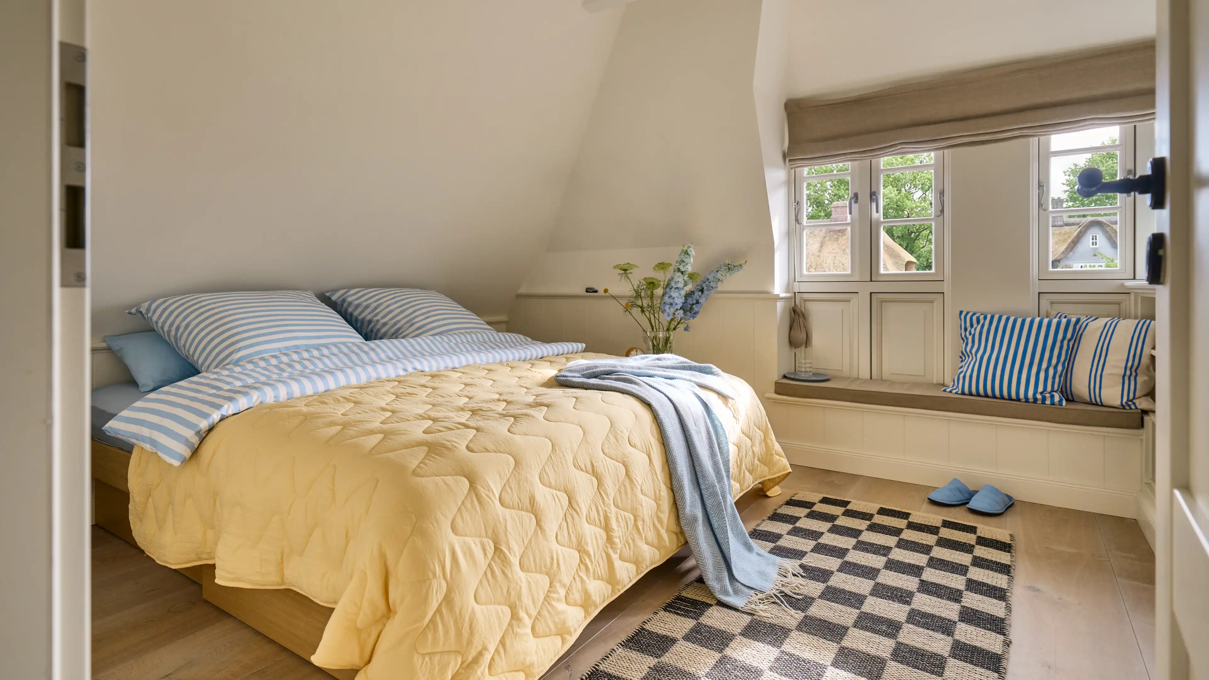 Das Hauptschlafzimmer mit goßem King-Size Bett in pastell blau und gelb gehaltenden Akzenten. Neben dem Bett, ist eine kleine Sitznische direkt vor den Fenstern zu sehen.