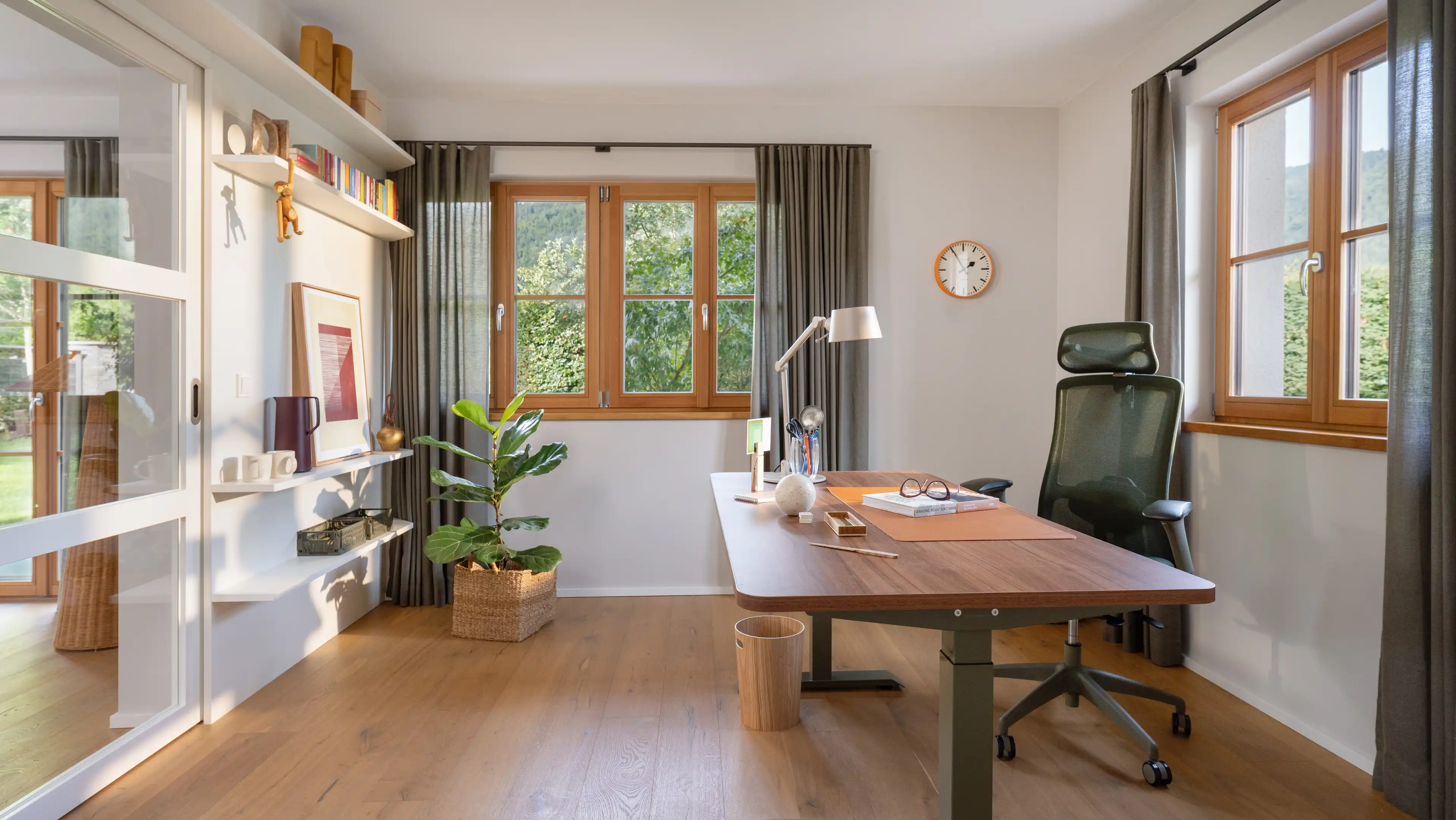 Arbeitszimmer mit dunklem Schreibtisch auf hoehenverstellbarem Gestell, schwarzem ergonomischen Buerostuhl und Wandregalen mit Buechern vor Fenstern mit Gartenblick.