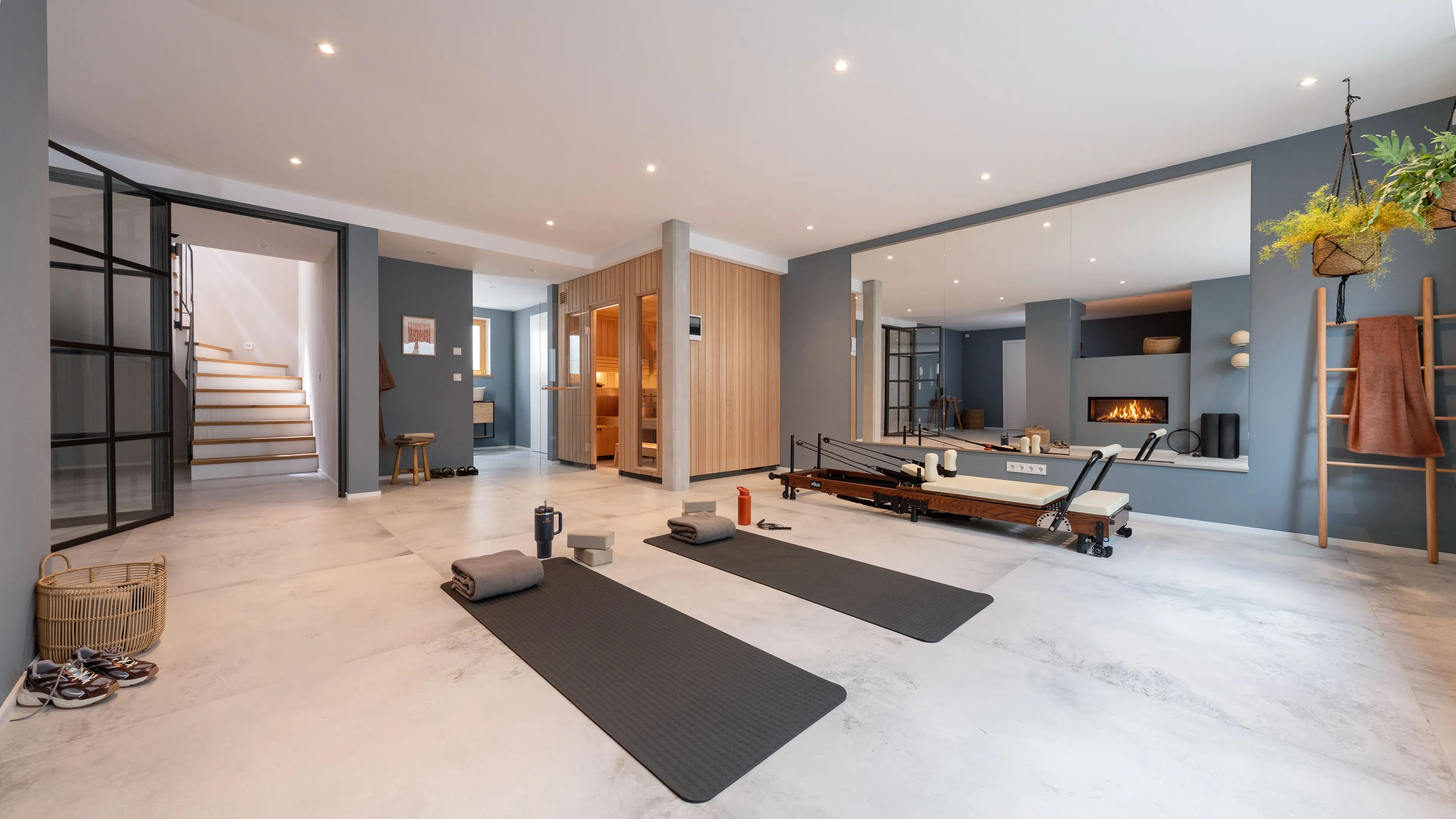 Fitnessraum mit Betonoptik-Boden, zwei schwarzen Yogamatten, einem hoelzernen Pilates-Reformer und einer Spiegelwand, die Kamin und Treppenaufgang reflektiert.