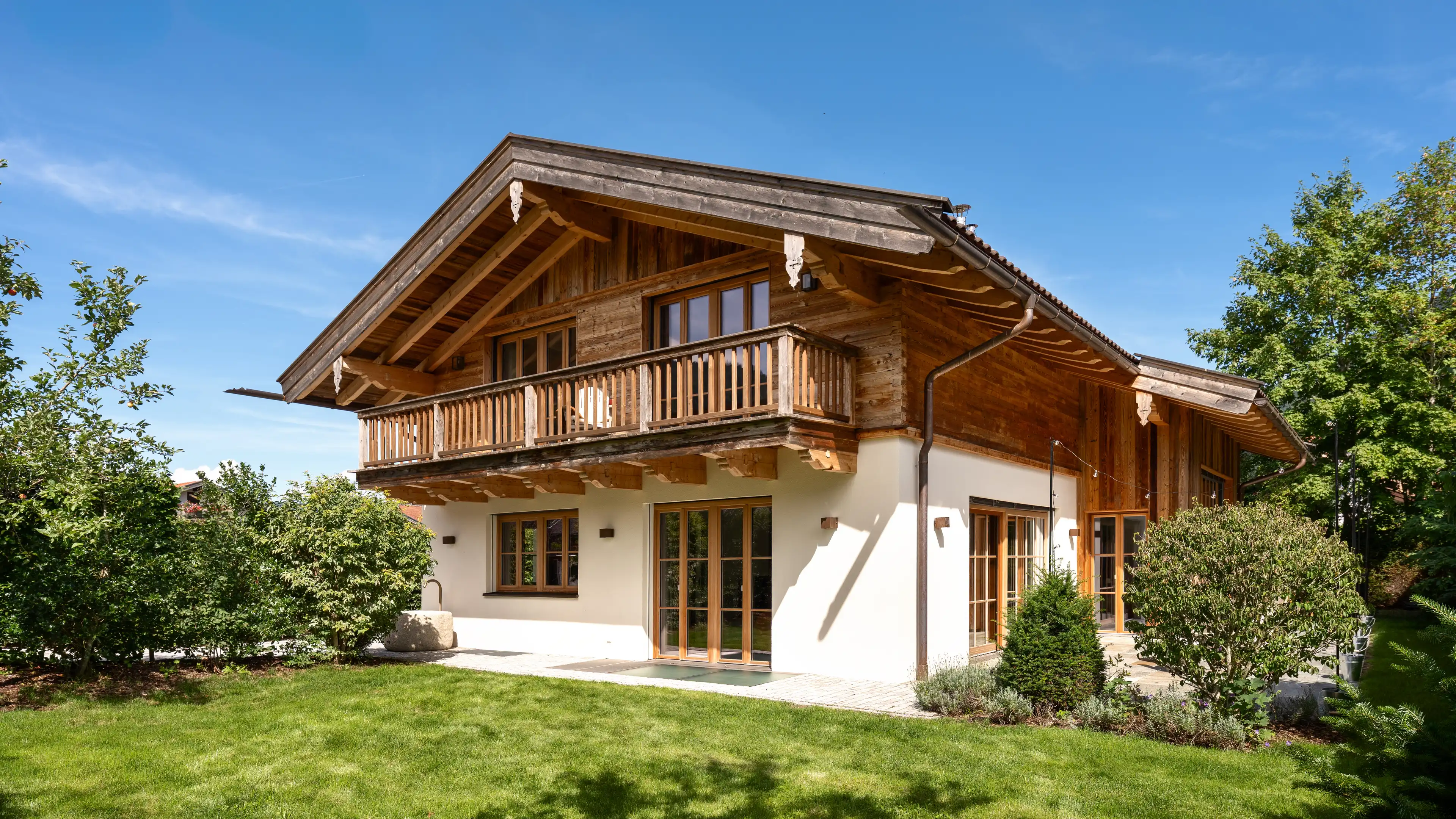 Frontansicht eines zweistöckigen Landhauses mit dunkler Holzfassade und auskragendem Giebeldach im Obergeschoss, weiß verputztem Erdgeschoss mit Holzfenstern und Terrassentüren, umgeben von grünem Rasen, Hecken und Bäumen unter blauem Himmel.
