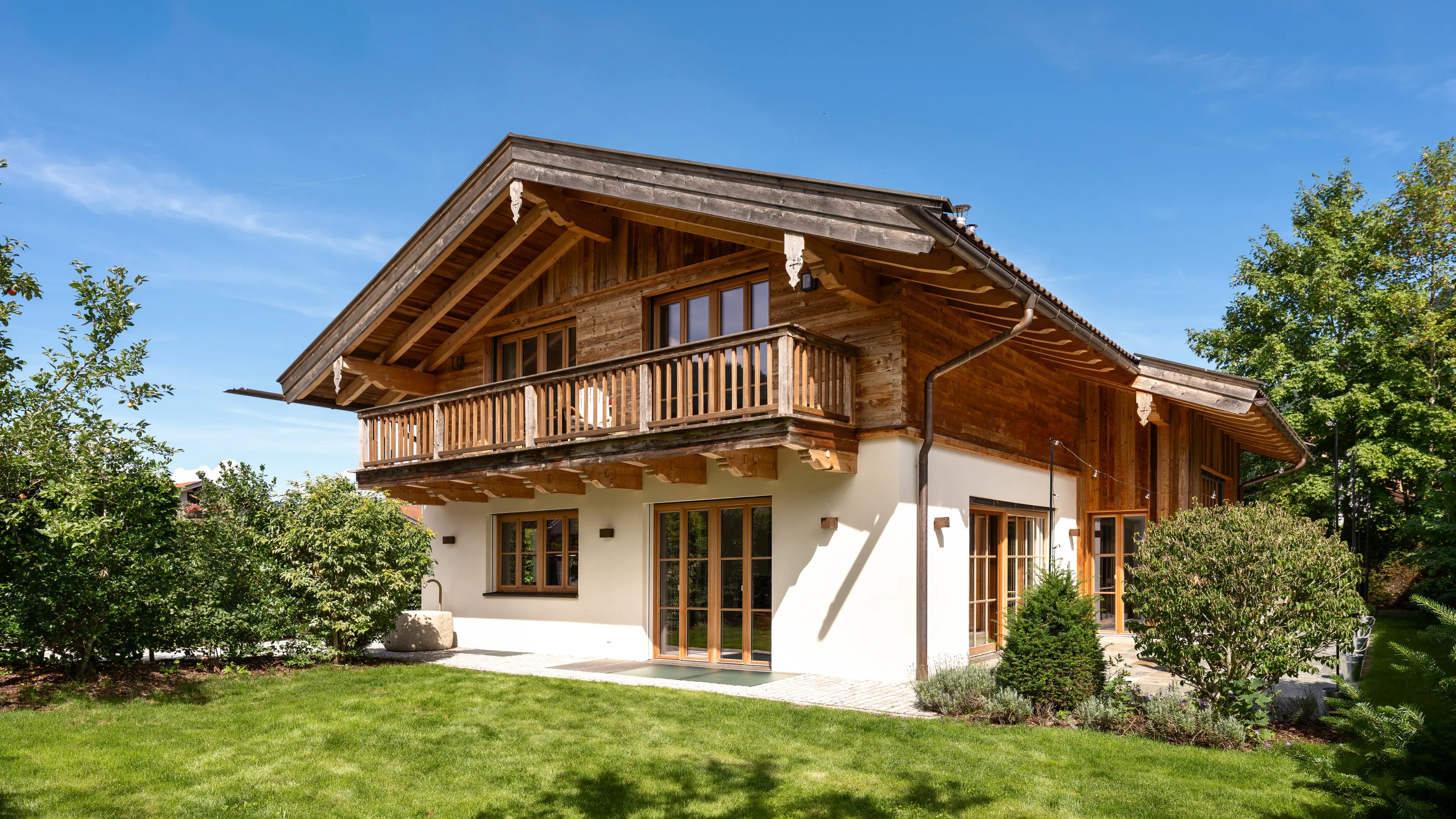 Frontansicht eines zweistöckigen Landhauses mit dunkler Holzfassade und auskragendem Giebeldach im Obergeschoss, weiß verputztem Erdgeschoss mit Holzfenstern und Terrassentüren, umgeben von grünem Rasen, Hecken und Bäumen unter blauem Himmel.