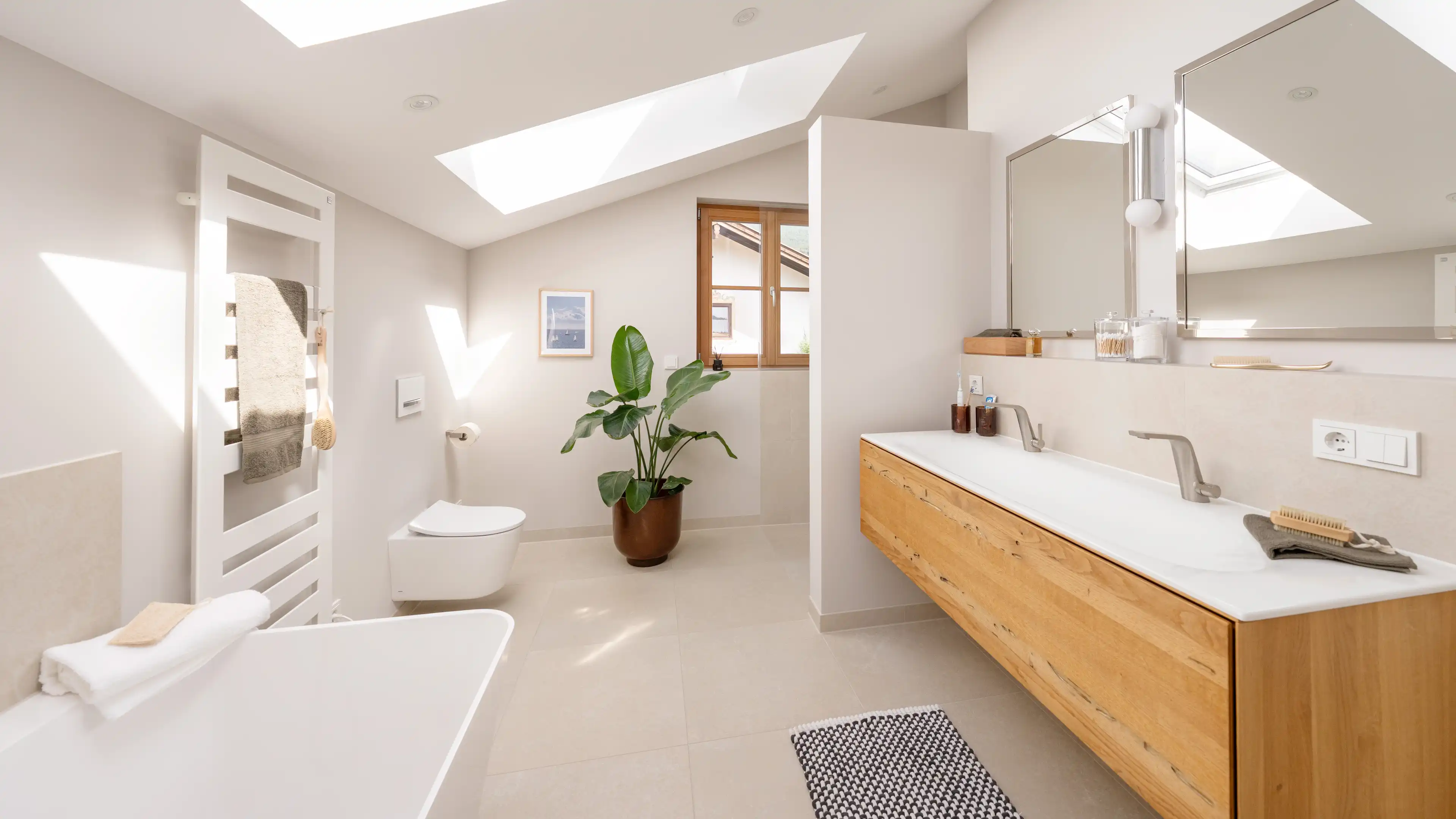 Badezimmer mit Dachfenster, freistehender weisser Badewanne, wandhaengendem WC, grosser Gruenpflanze und einem hellen Doppelwaschtisch aus Holz mit zwei Spiegeln.