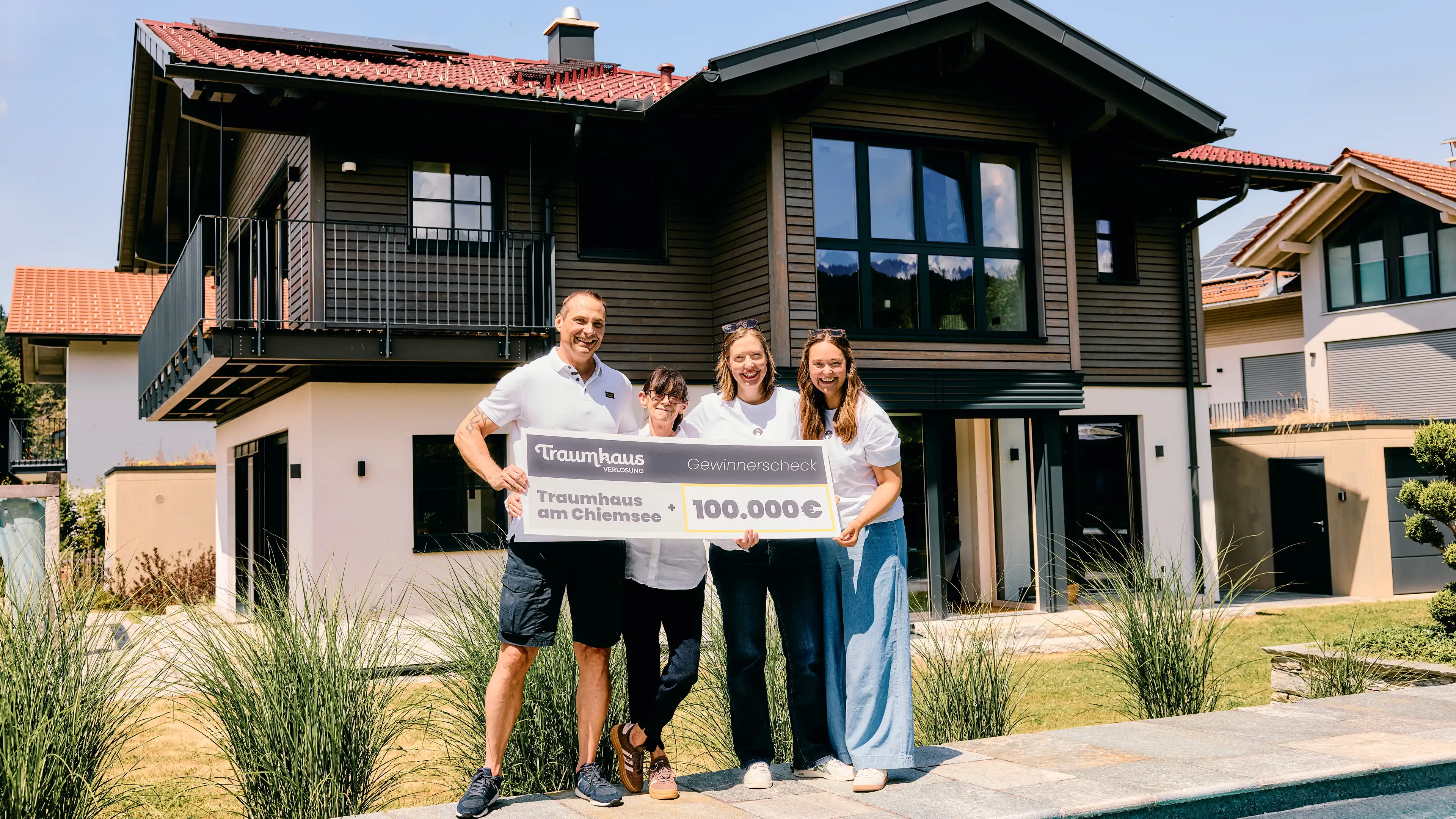 Michael, der Gewinner vom Traumhaus am Chiemsee mit seiner Frau und Fredi und Larissa von der Traumhausverlosung mit einem Check in Höhe von 100.000 € vor dem Traumhaus am Chiemsee