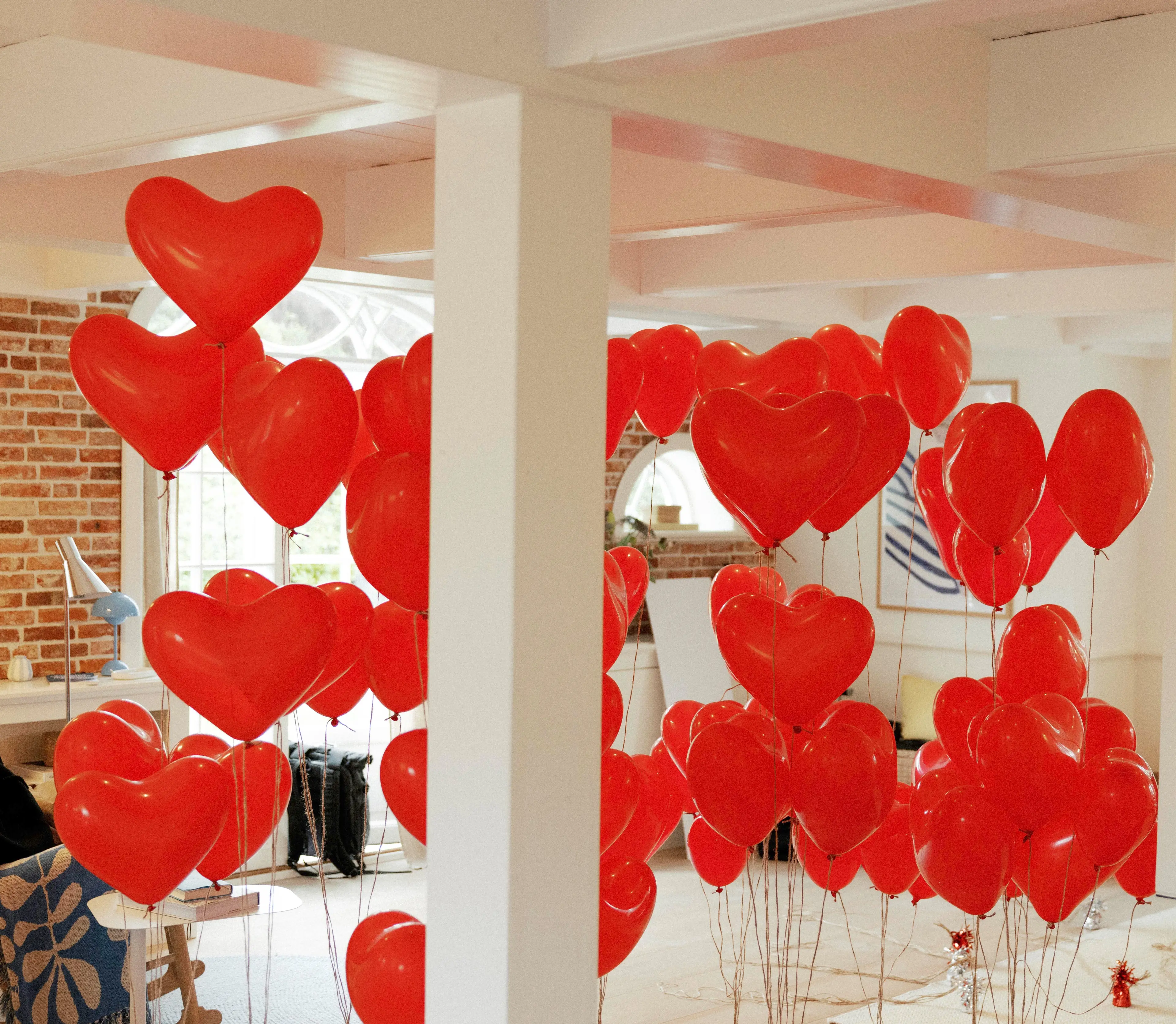 Room filled with red heart-shaped balloons tied with strings, set against a modern interior with white and brick walls.
